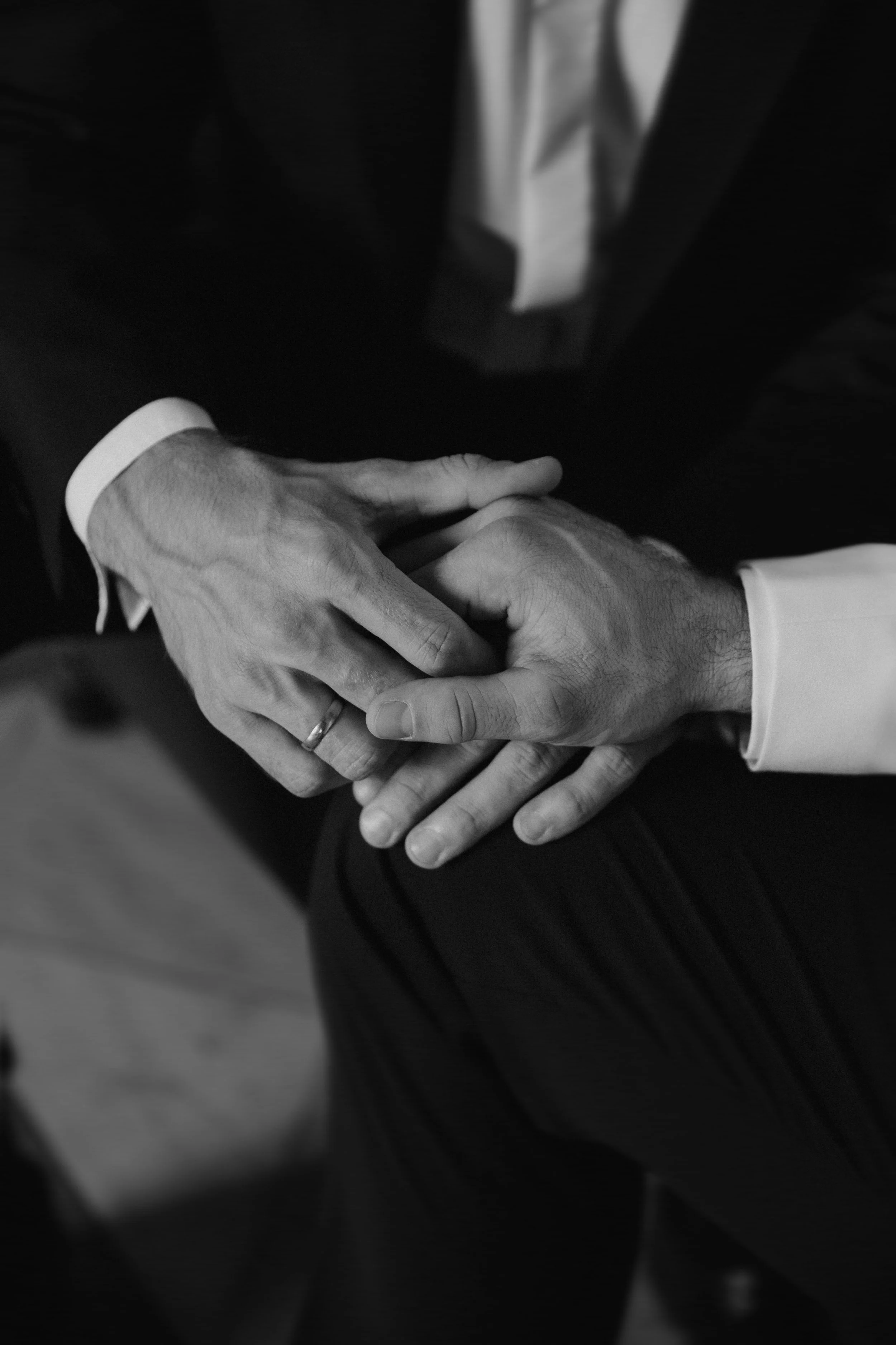 Zwei Männer in formeller Kleidung halten sich beim Sitzend an den Händen, eines der Männer trägt einen Ehering.