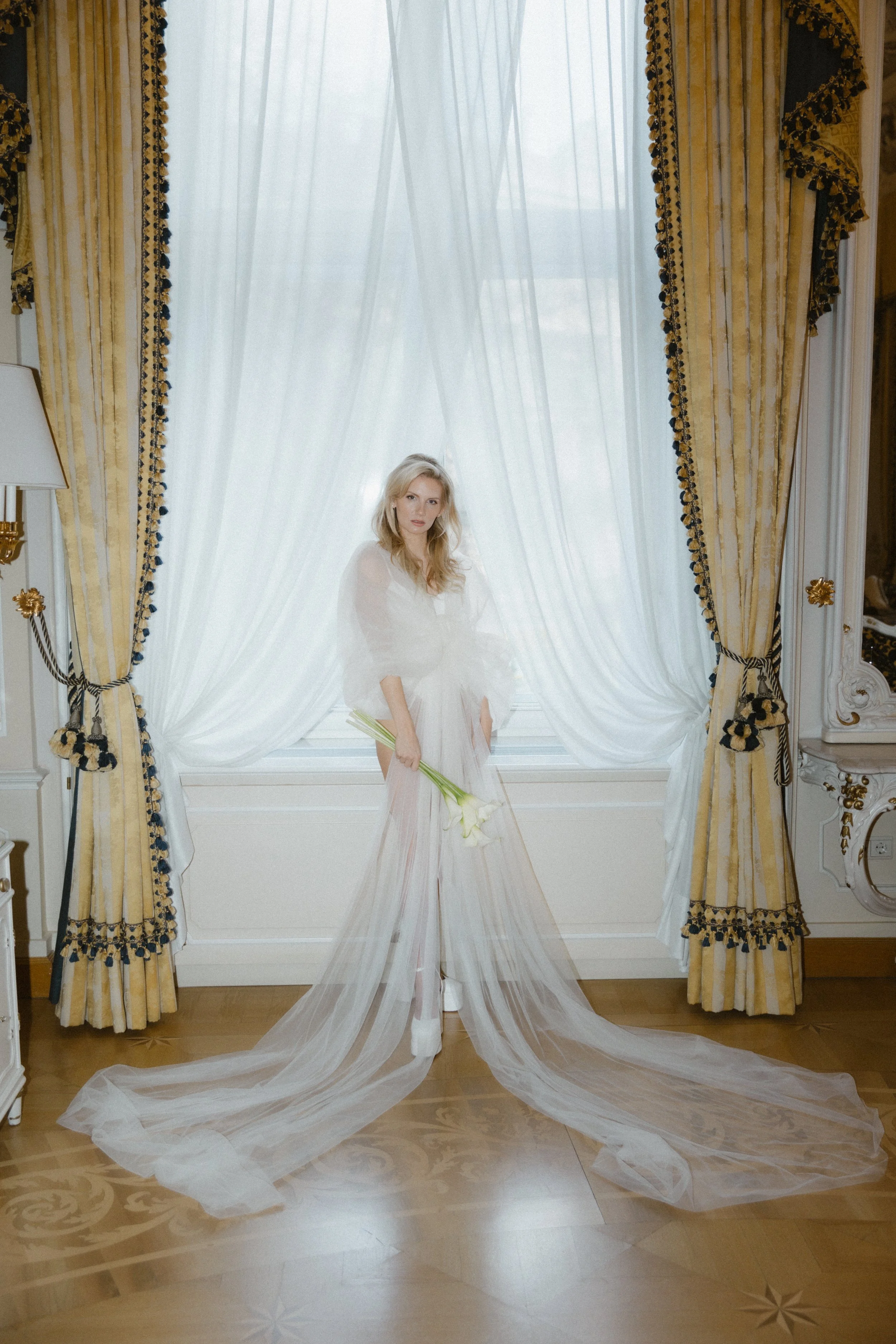 Frau in Brautkleid mit Blumen im Raum mit goldenen Vorhängen und weißen Gardinen vor einem Fenster.
