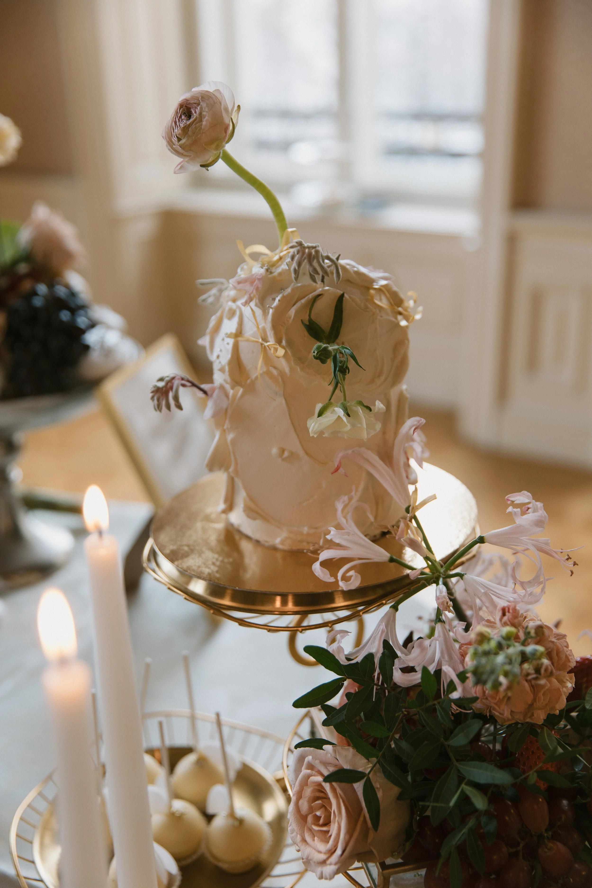 Eine zeremonielle Torte mit Blumen, auf einem goldenen Kuchenständer, umgeben von Kerzen und Blumenarrangements, im Hintergrund eine helle Fensterfront.
