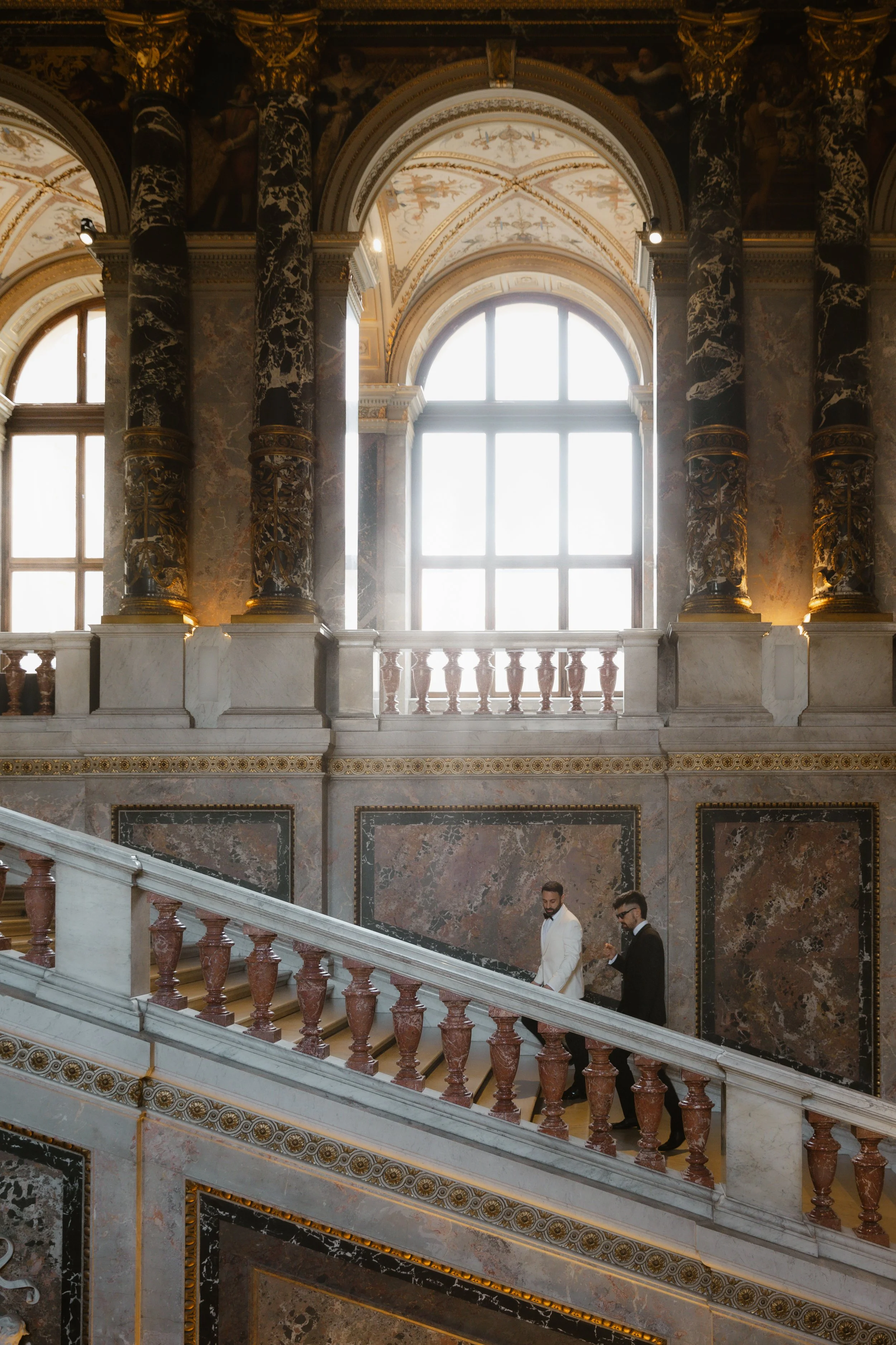 Zwei Männer in formeller Kleidung gehen die Treppe in einem prachtvollen, historischen Gebäude mit großen Fenstern und kunstvoll verzierten Wänden hoch.