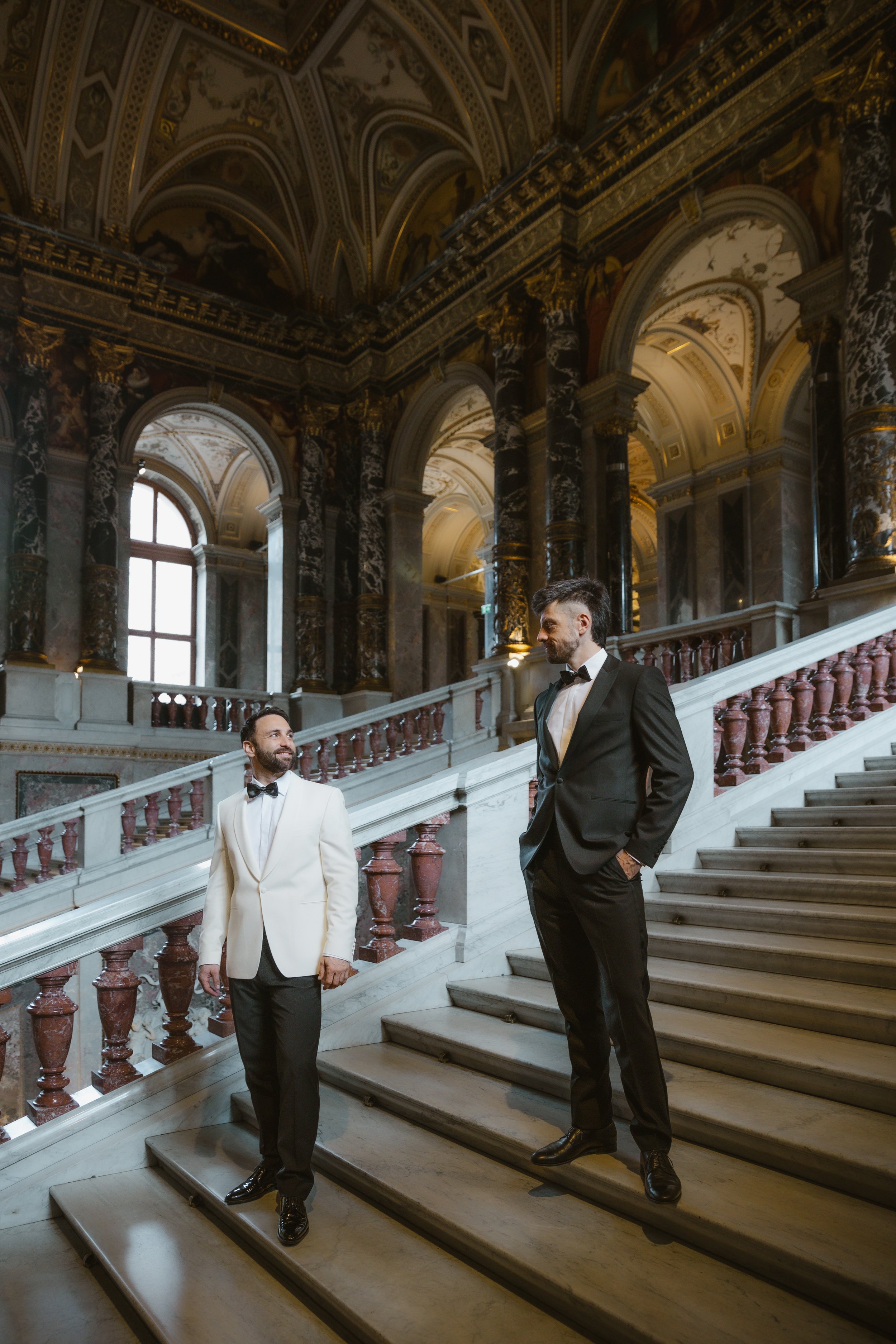 Zwei Männer in formeller Kleidung stehen auf einer prunkvollen Marmortreppe in einer barocken Halle mit hohen Fenstern und goldverzierten Decken.