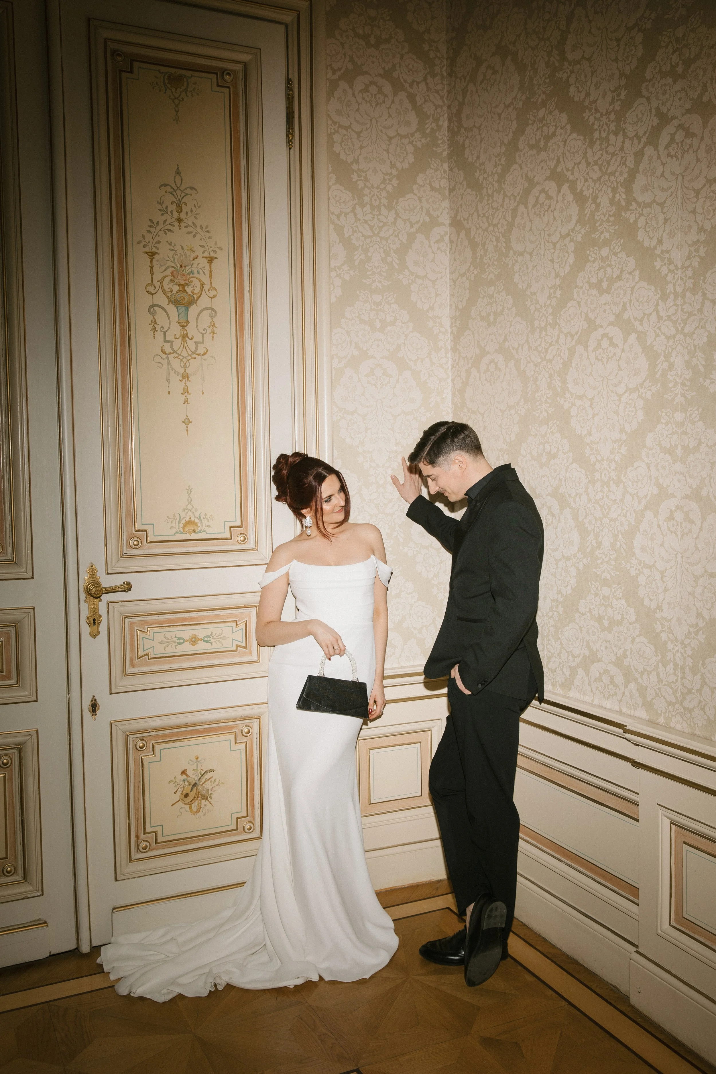 Ein Brautpaar in eleganten Kleidern posiert in einem Raum mit reichen, barocken Wanddekorationen und Holzfußboden.