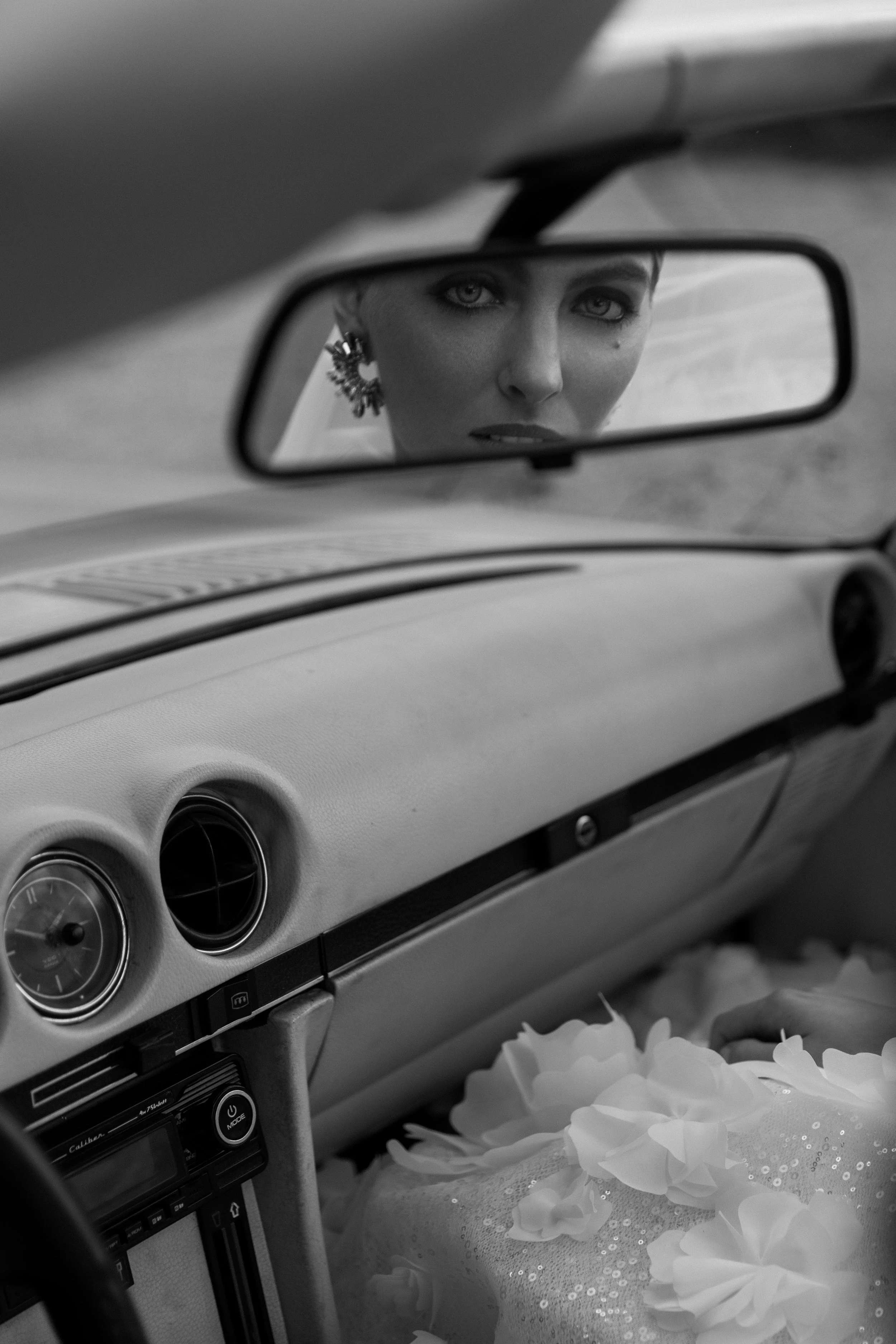 Schwarz-Weiß-Foto eines Autoinnenraums mit Spiegel, der eine Frau mit kurzer Frisur, Ohrringen und Make-up zeigt.