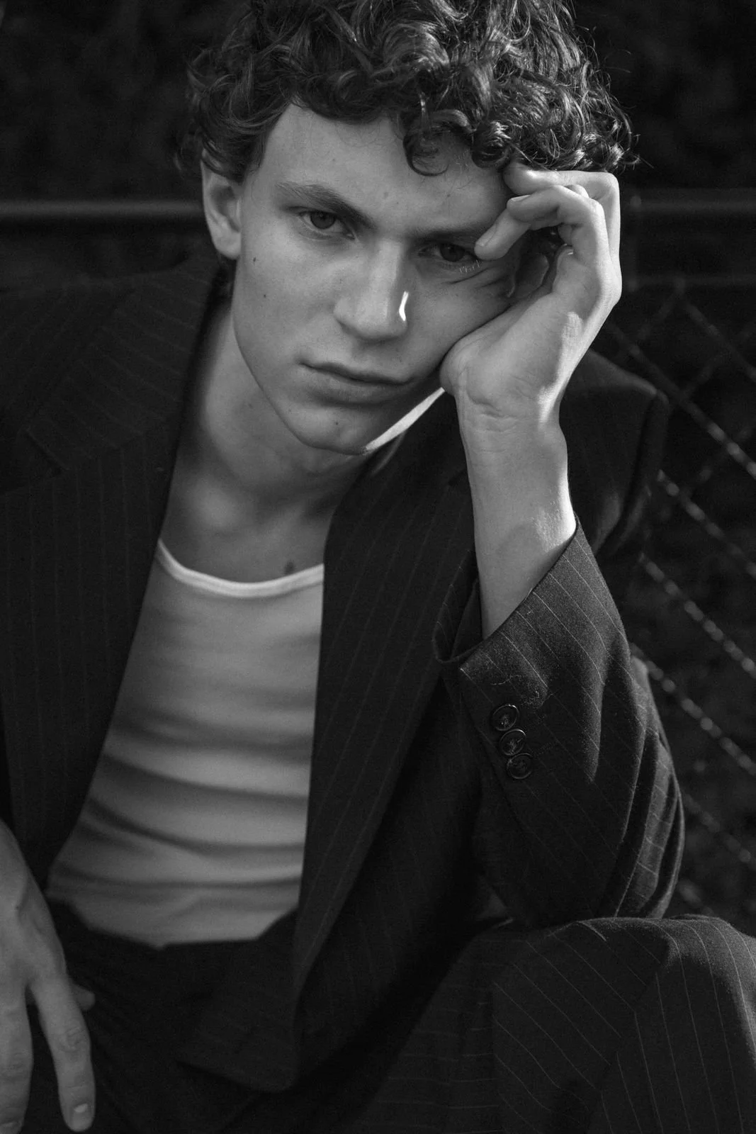 Schwarz-weiß-Foto eines jungen Mannes mit lockigem Haar, der eine nachdenkliche Pose einnimmt, sein Kinn mit der Hand stützt und direkt in die Kamera schaut.
