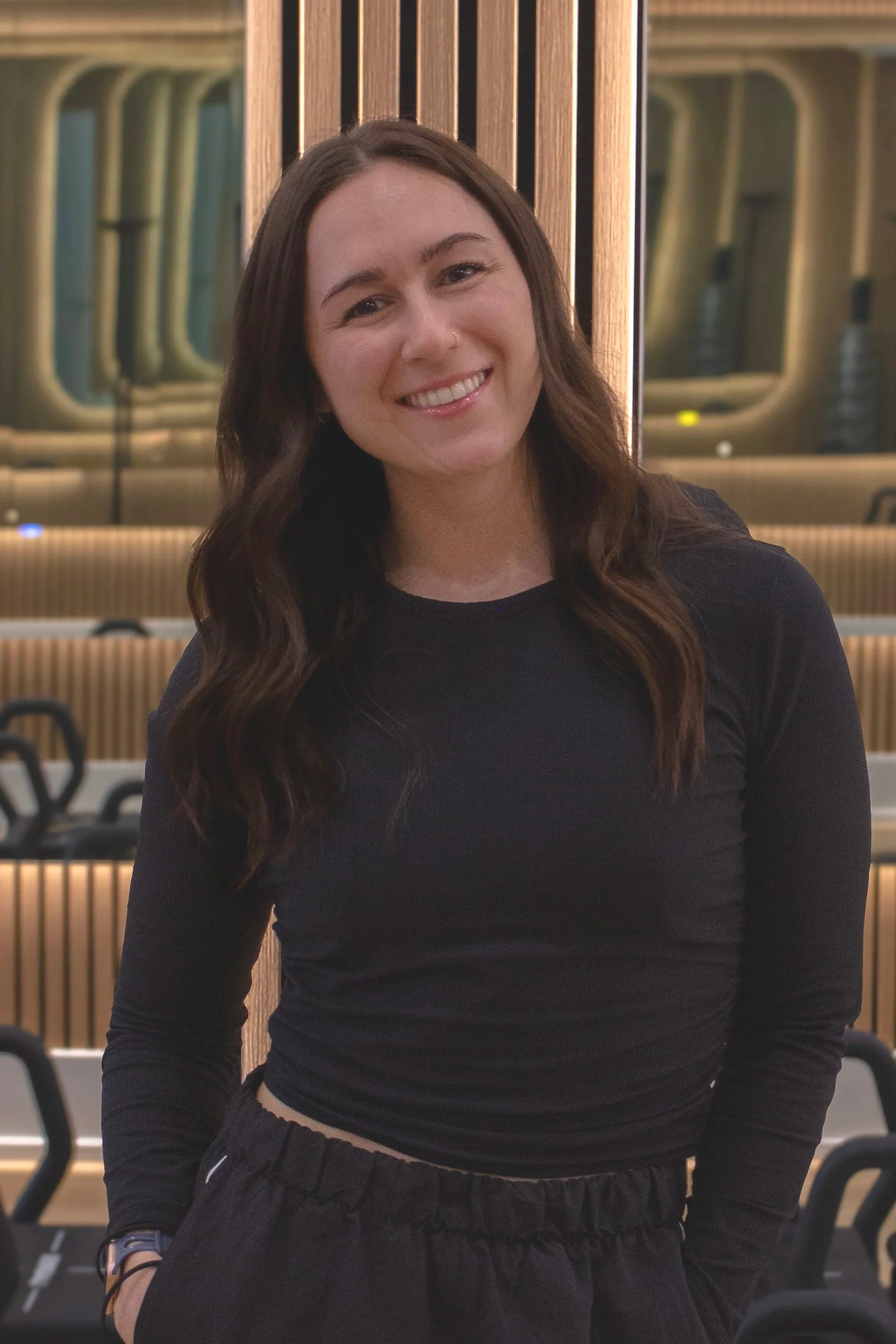 A young woman with long wavy brown hair and a nose piercing smiling at the camera, dressed in a black long-sleeve shirt, standing in an indoor setting with wooden panels and chairs in the background.