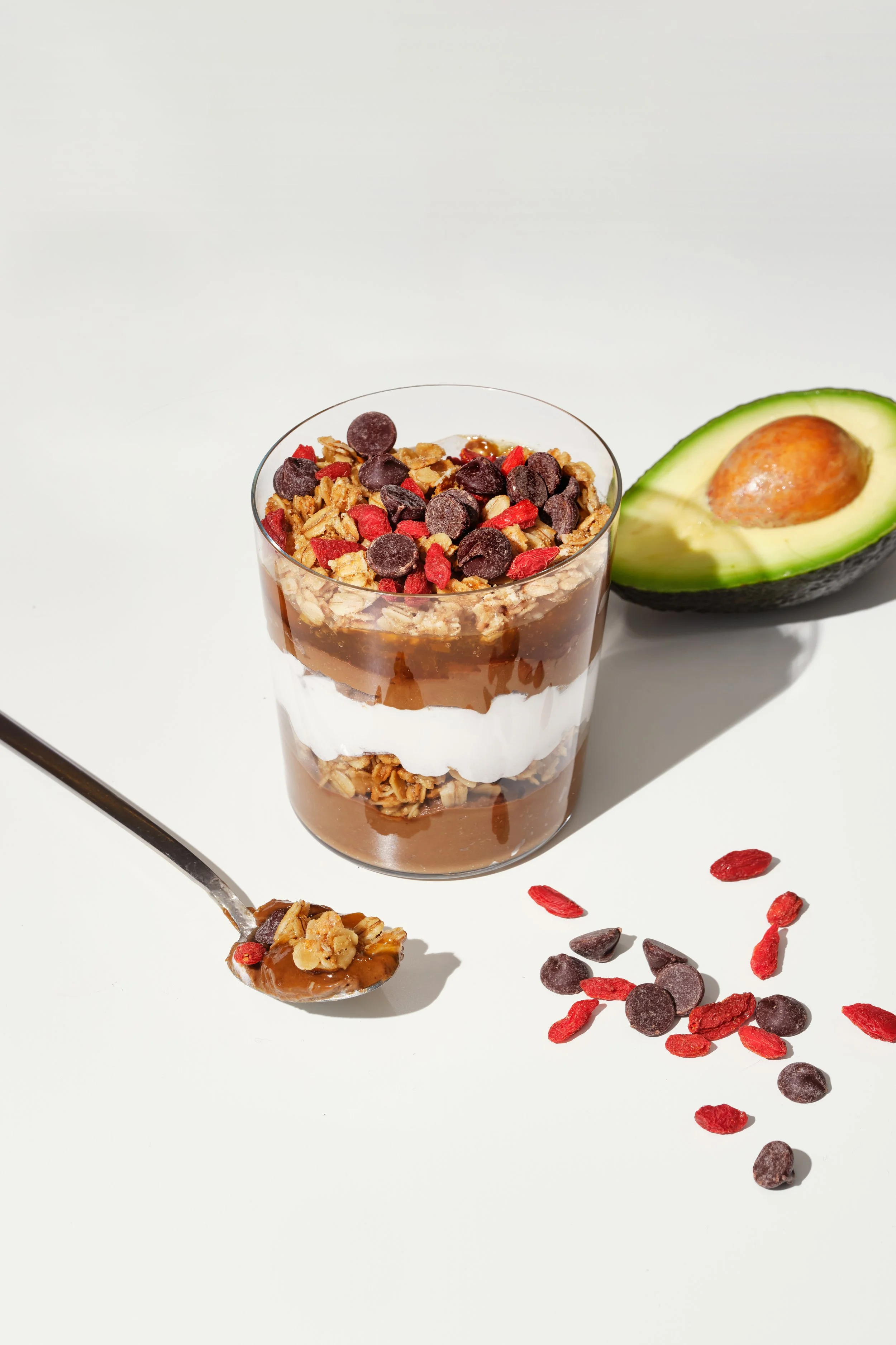 A layered chocolate parfait with granola, whipped cream, and chocolate sauce in a glass, garnished with chocolate chips and goji berries; a spoonful with granola and goji berries, half an avocado, and scattered goji berries and chocolate chips on a white surface.