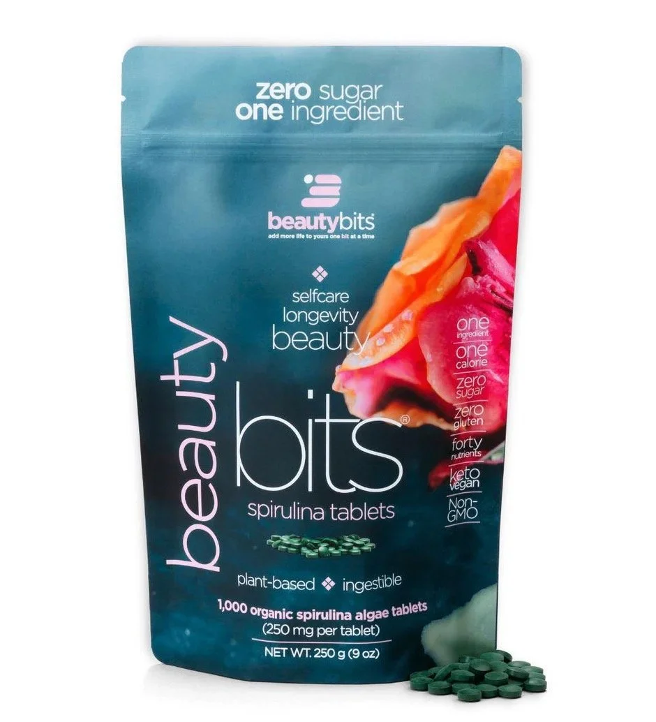 A teal-colored resealable pouch of Beauty Bits spirulina tablets with product name and features, partially open with green tablets spilling out, and a vibrant, abstract background of pink and orange flowers.