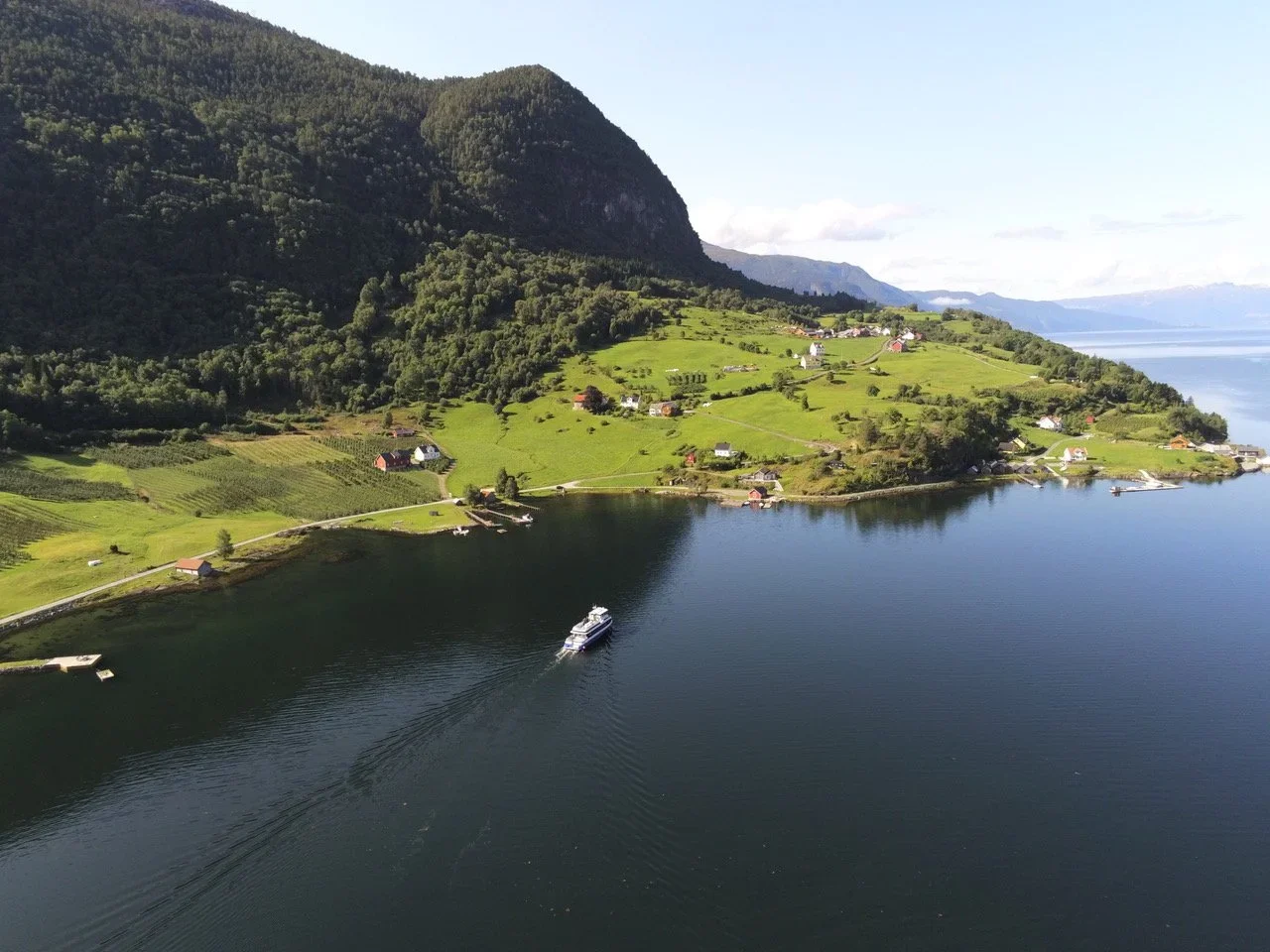 A scenic view of a coastal area with green hills, scattered houses, and a boat on calm water, with mountains in the background under a clear sky.