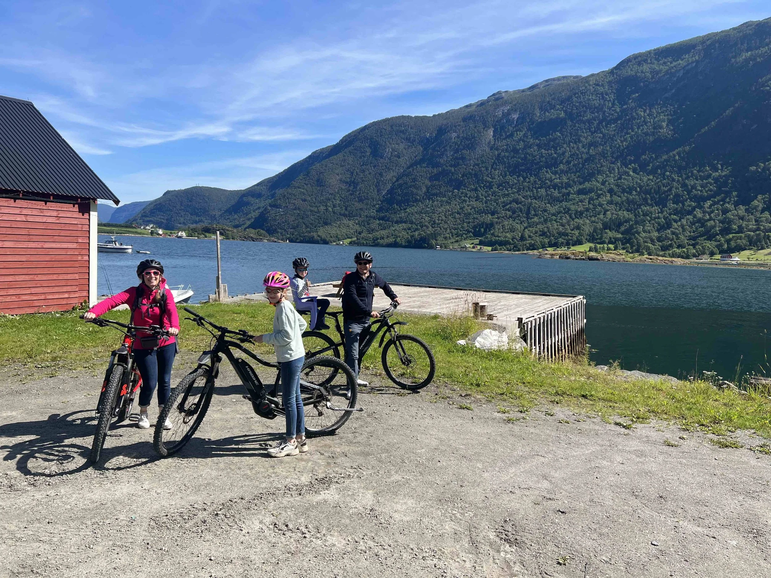 Four people in helmets with bicycles near water and green mountains under a blue sky