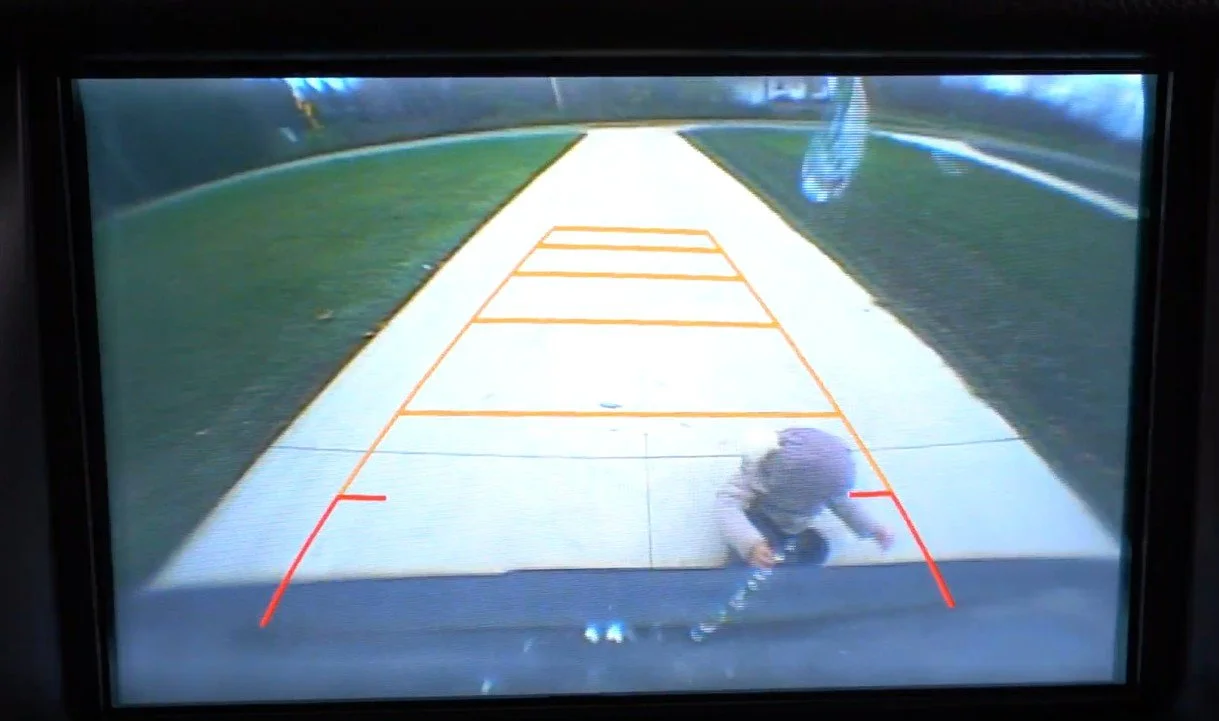 A security camera view of a driveway with a person in dark clothing squatting near the bottom of the screen, with parking guidelines overlaid.