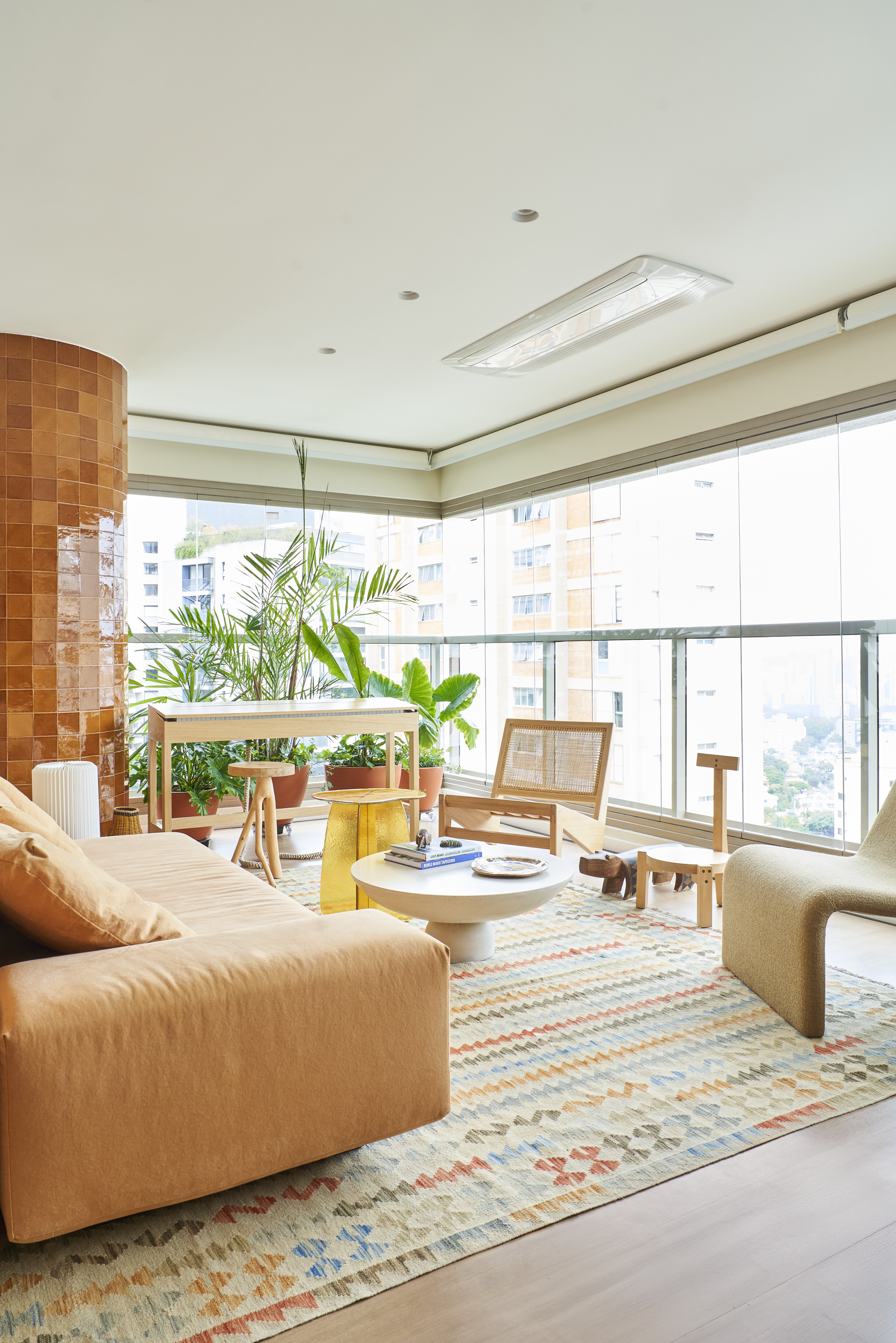 Sala de estar moderna com sofá de veludo laranja, mesa de centro branca, poltrona de rattan decorativa, tapete colorido e muitas plantas em vasos, janelas amplas com vista para edifícios