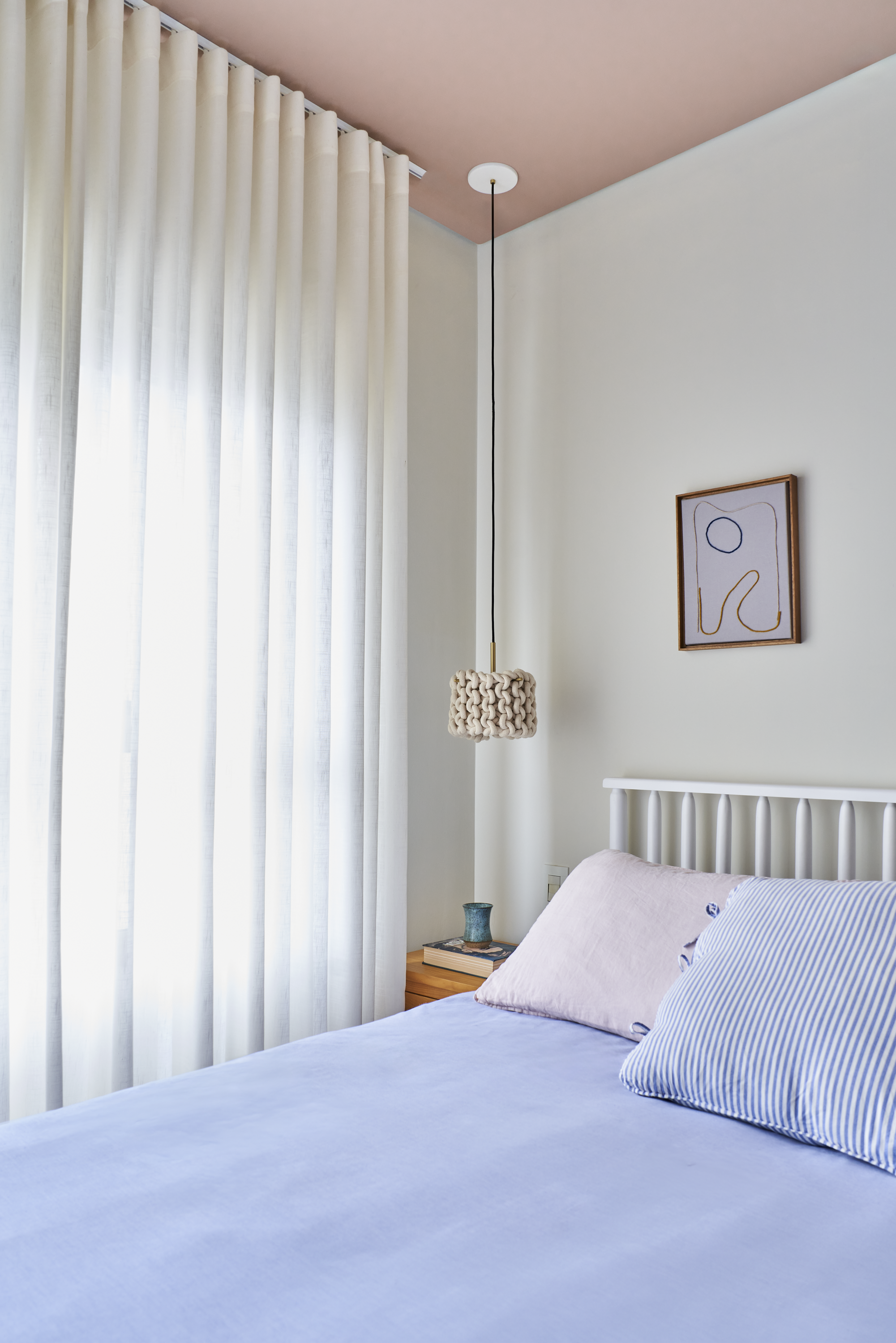 Quarto com cortinas brancas, parede branca, rede de dormir pendurada no teto com lâmpada de cabeceira de nadar, quadro minimalista na parede, cabeceira branca e roupas de cama em tons pastel.