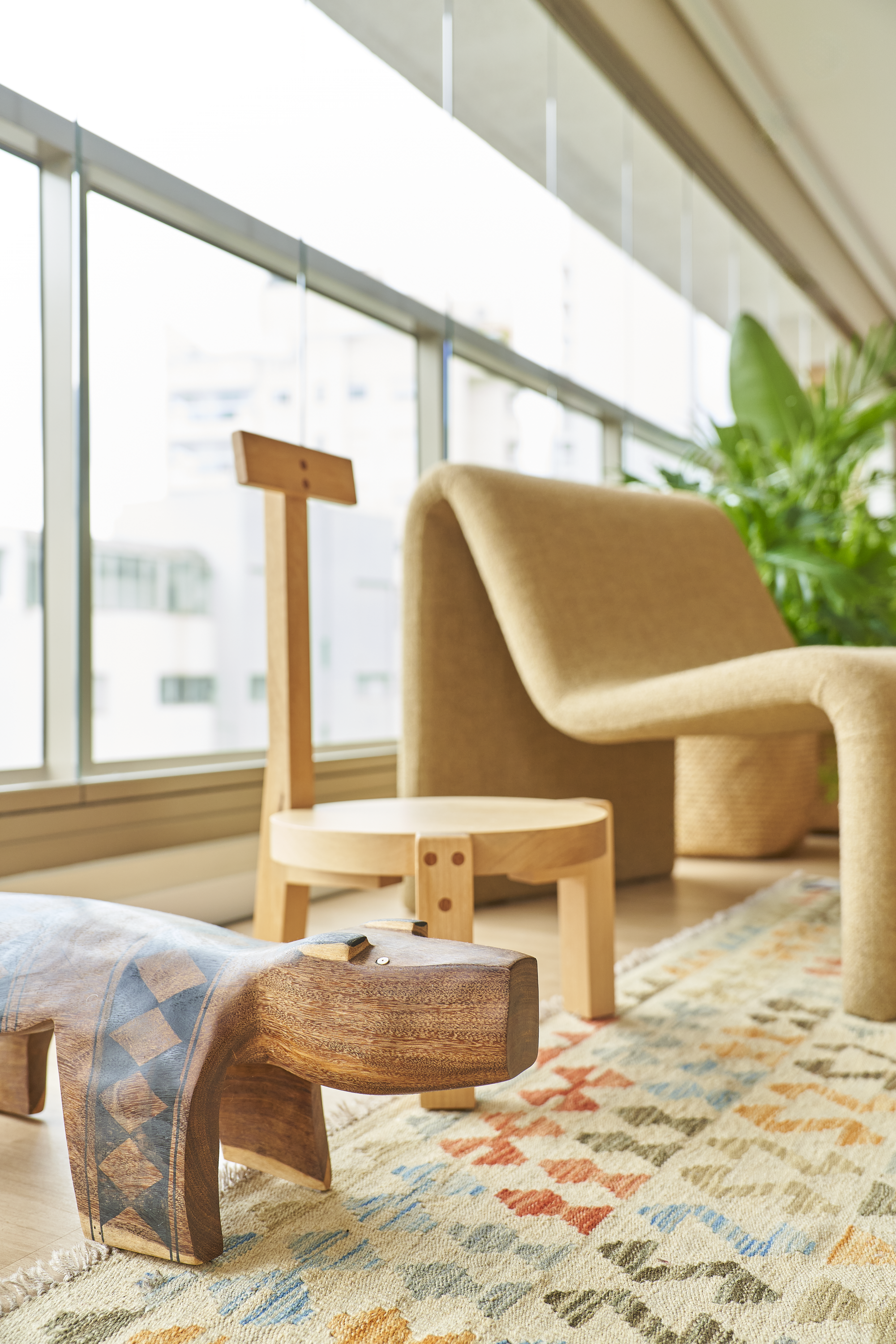 Sala de estar com sofá bege, poltrona de madeira, tapete colorido e muitas plantas, com janelas grandes de vidro ao fundo.