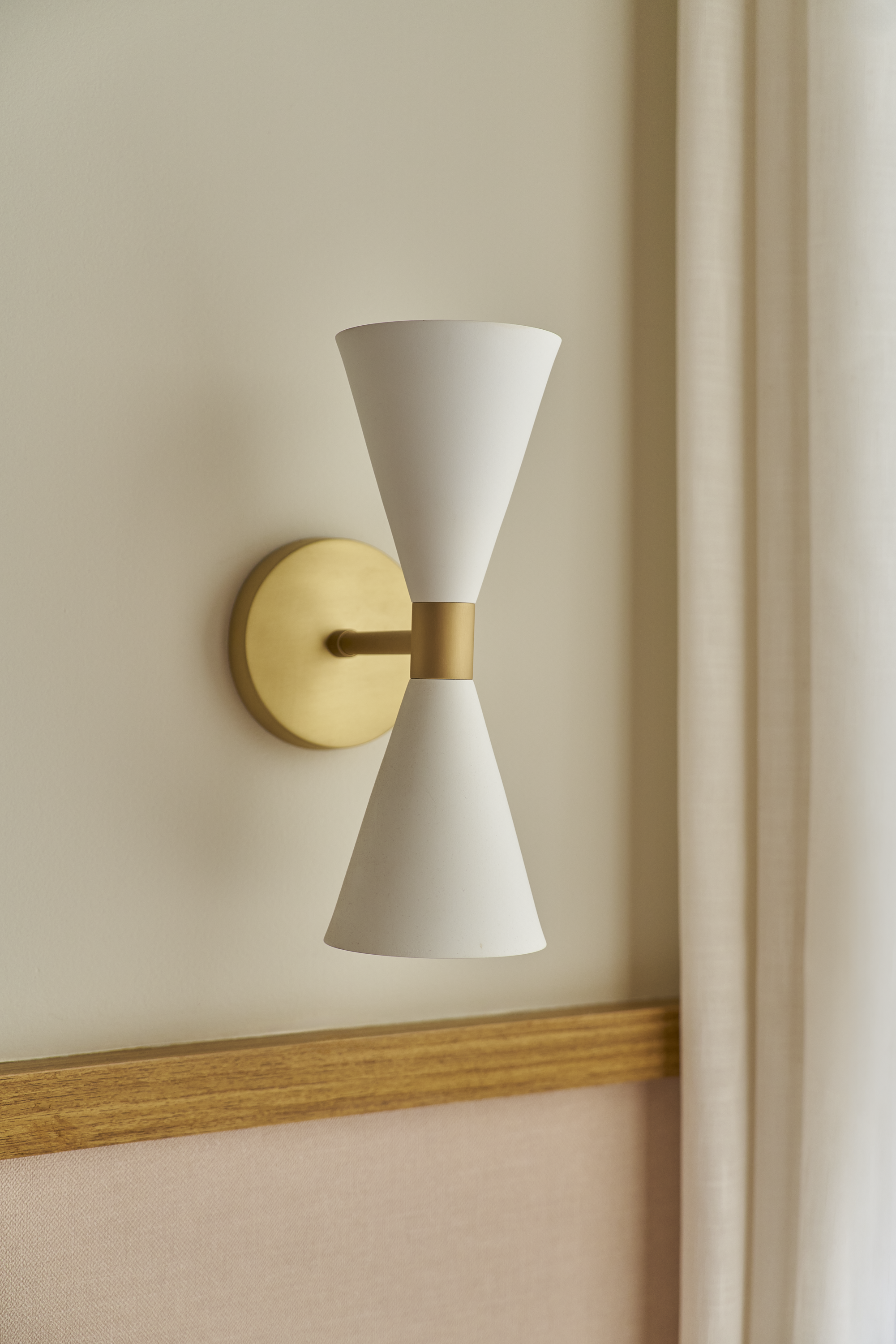 Luminária de parede moderna com dois quartos cônicos brancos unidos por uma conexão dourada, instalada em parede clara ao lado de cortinas bege.