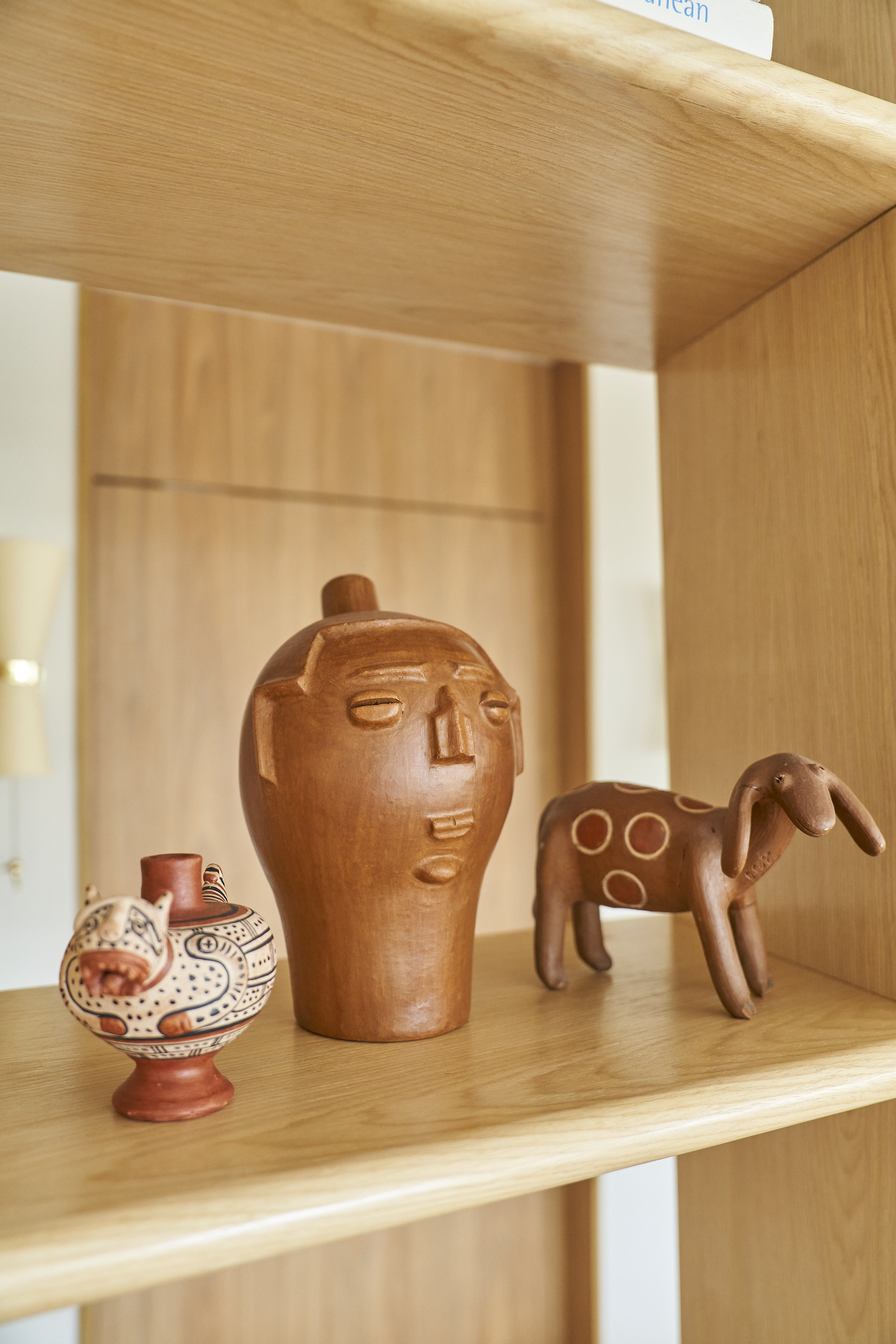 Três esculturas de cerâmica em uma prateleira de madeira: uma de forma de rosto humano com feições simples, uma de um animal com corpo arredondado e manchas circulares, e uma de um animal com corpo alongado e patas curtas.