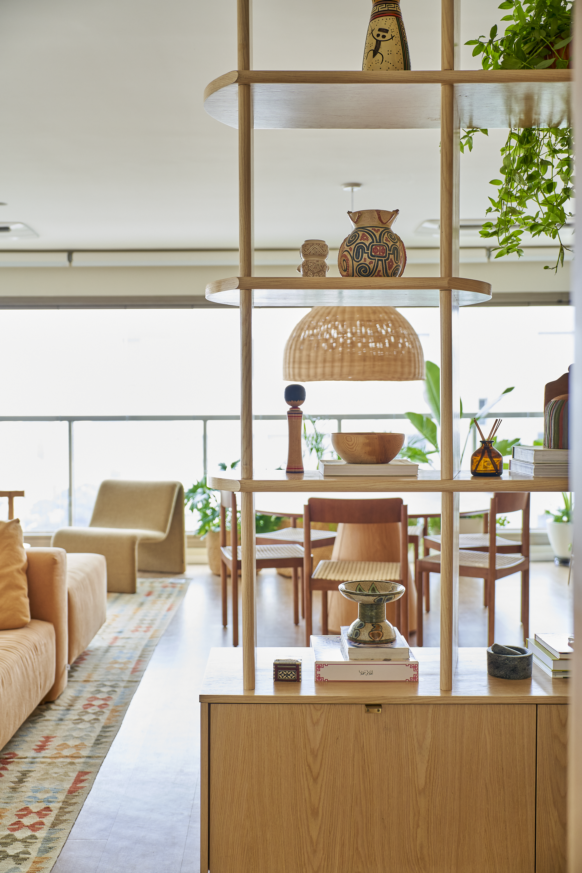 Estante de madeira decorada com vasos tradicionais, uma lâmpada de fibra natural, um recipiente cerâmico e livros, em um espaço com móveis de madeira, plantas e uma poltrona em fundo.