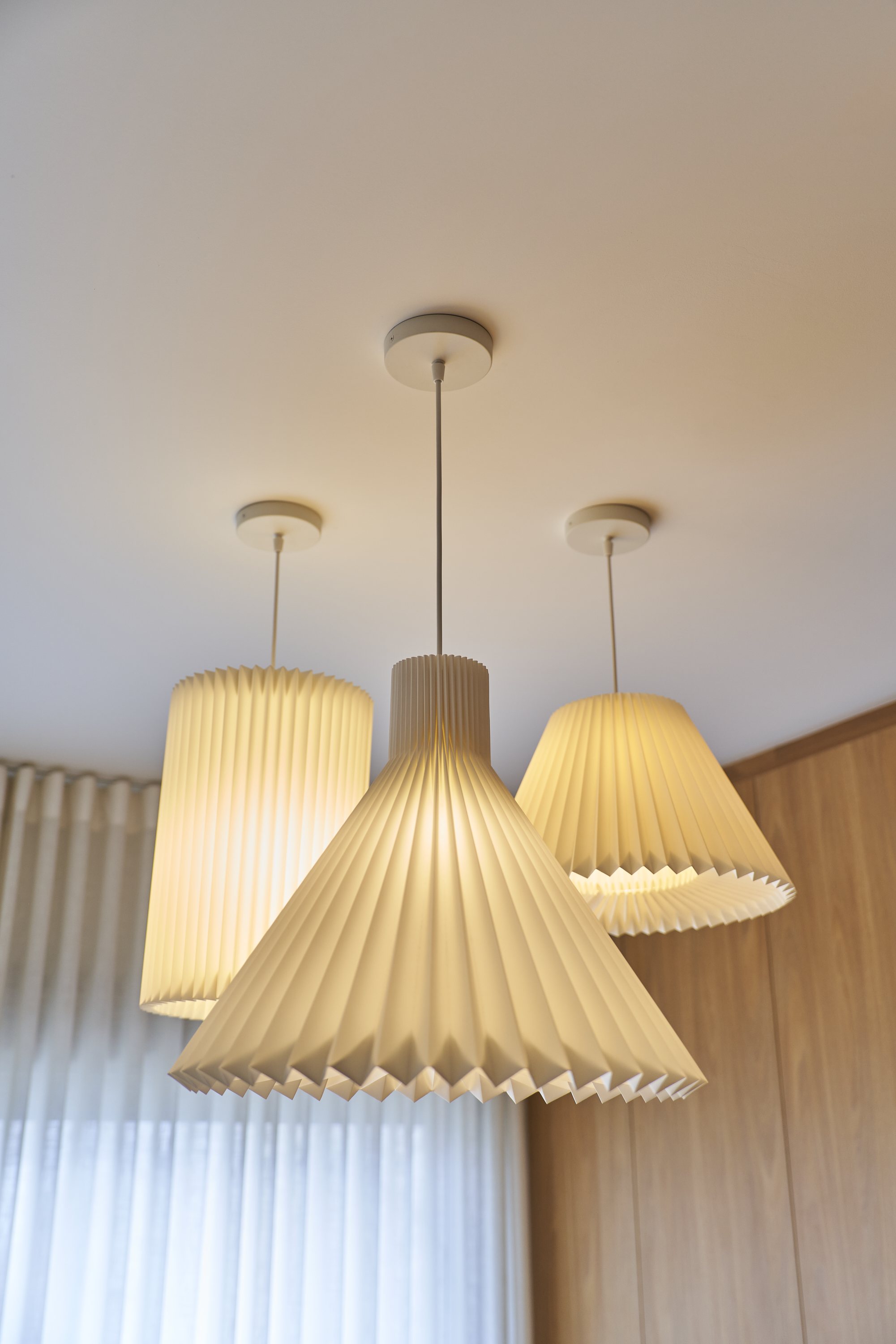 Lustre moderno com três luminárias de papel de tom bege, penduradas no teto de um ambiente com cortinas claras e parede de madeira.