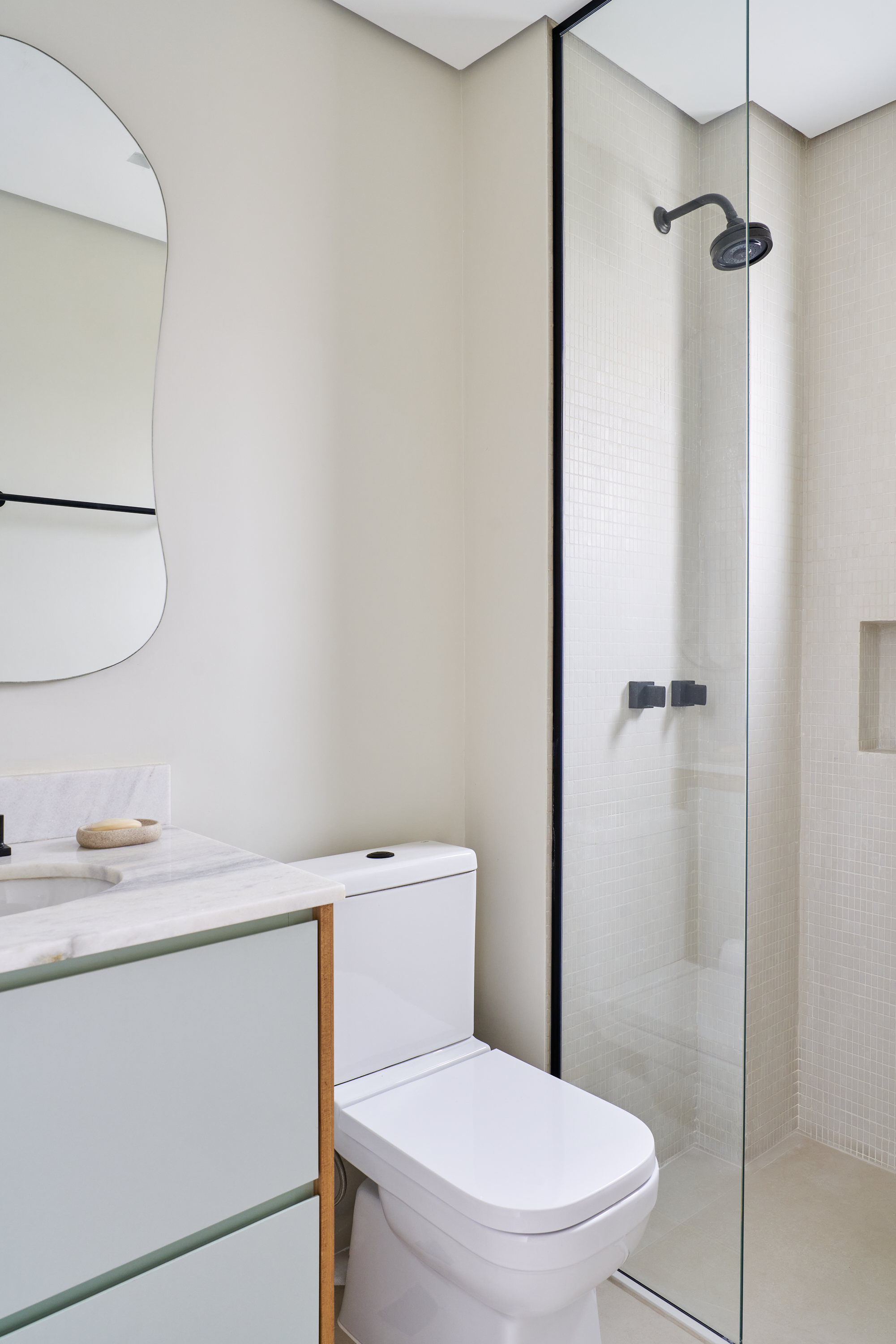 Banheiro com espelho de formato irregular, pia de mármore com sabonete, vaso sanitário e ducha de banho com vidro na parede.