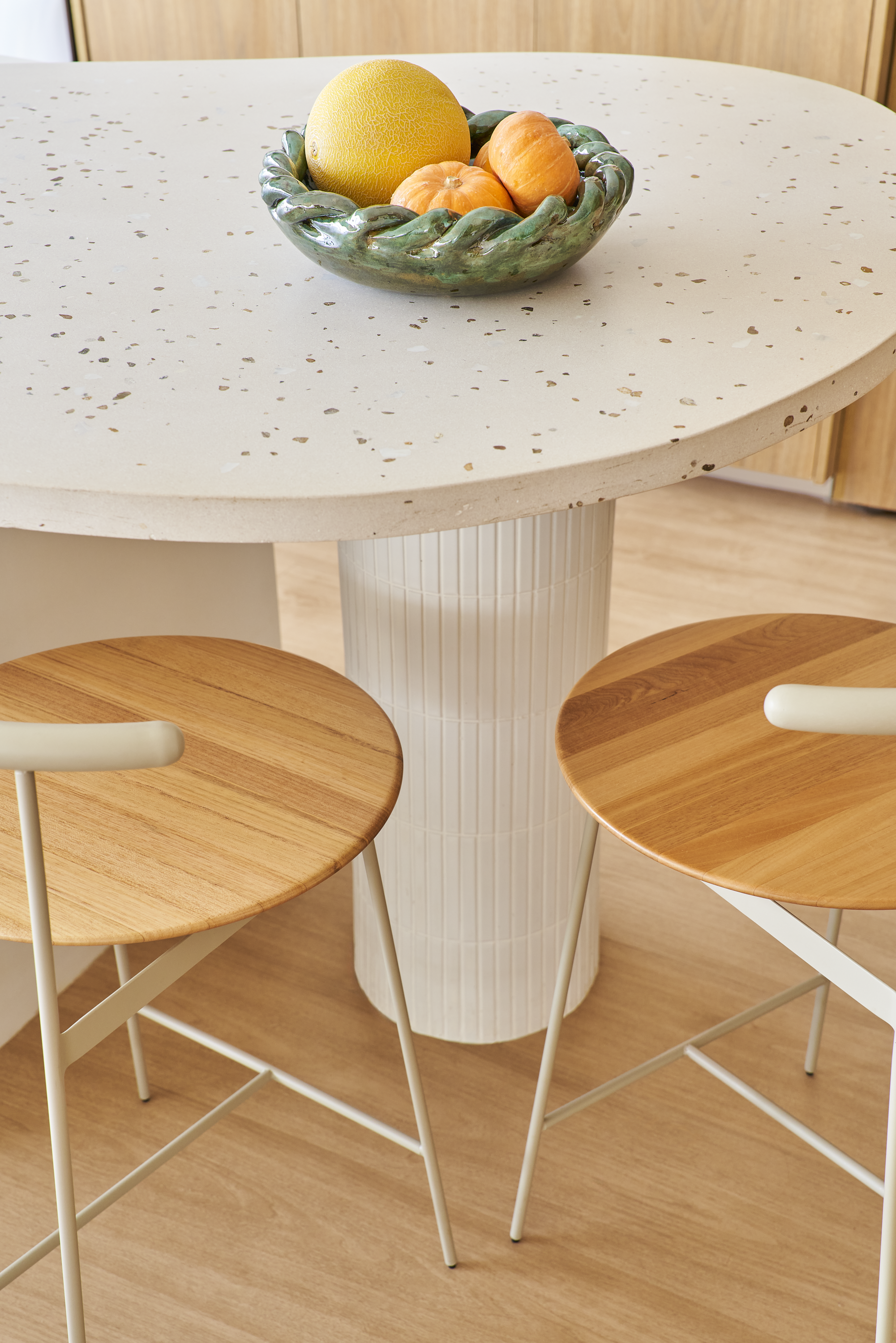 Mesa de vidro bege com base branca decorada com uma tigela de cerâmica com frutas, incluindo melão e tangerinas. Ao redor da mesa, duas cadeiras com assentos de madeira e estrutura branca.