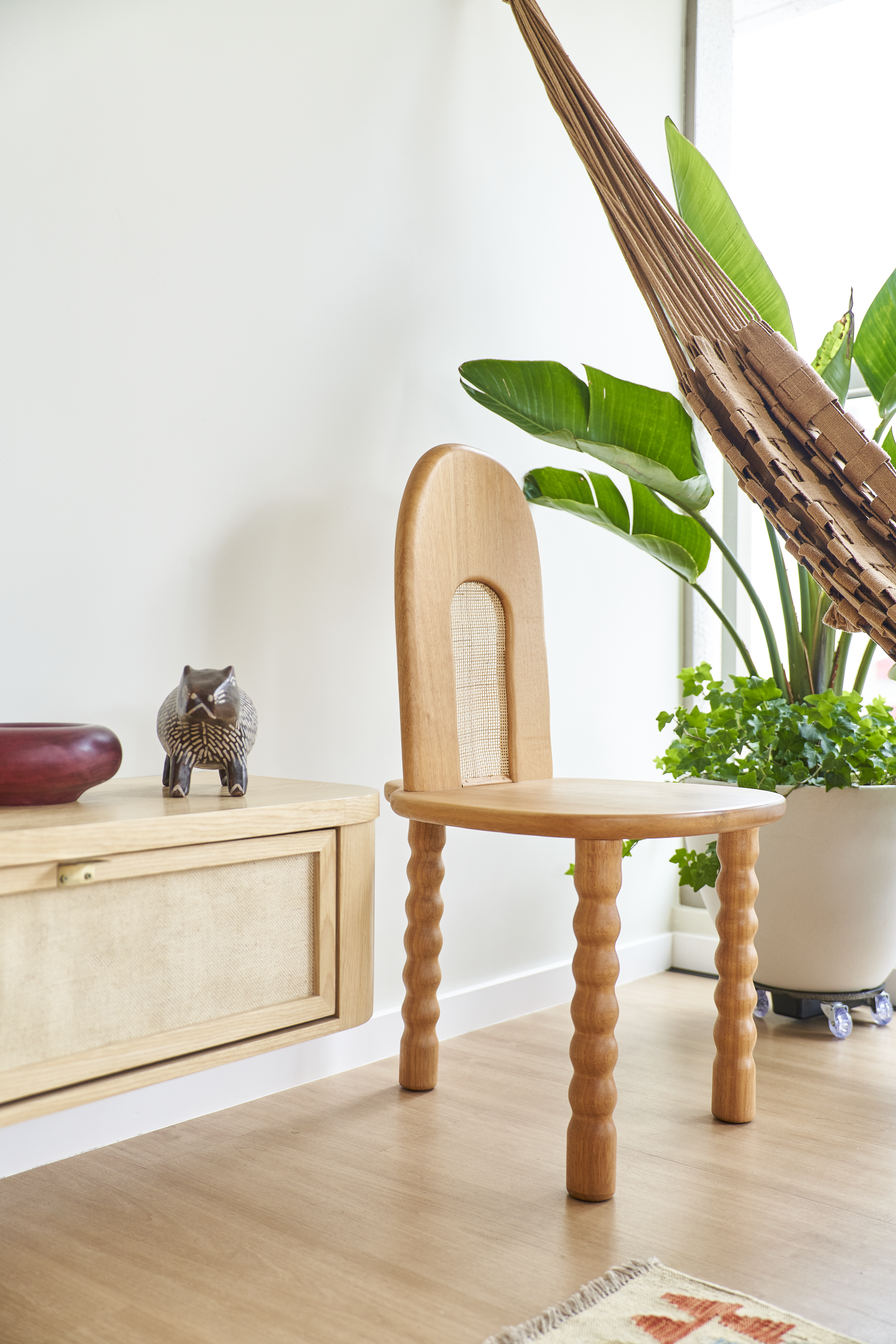 Cadeira de madeira com encosto de palha, perto de uma parede branca, ao lado de uma planta grande em vaso e uma mobília de madeira com uma cerejeira decorativa. Um pufe de palha com uma tapeçaria de tapete também está visível.