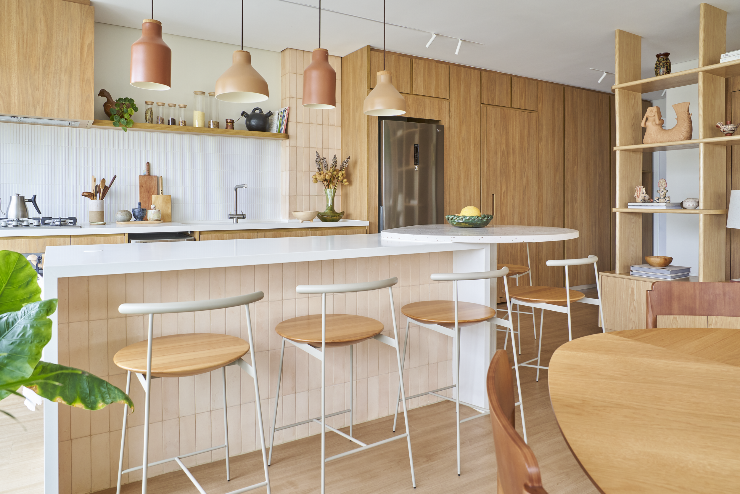 Cozinha moderna com balcão de madeira e cadeiras de madeira com assento branco, iluminação suspensa de cores neutras, armários de madeira, utensílios de cozinha e decoração de vasos e livros, ambiente bem iluminado e decorado com plantas.