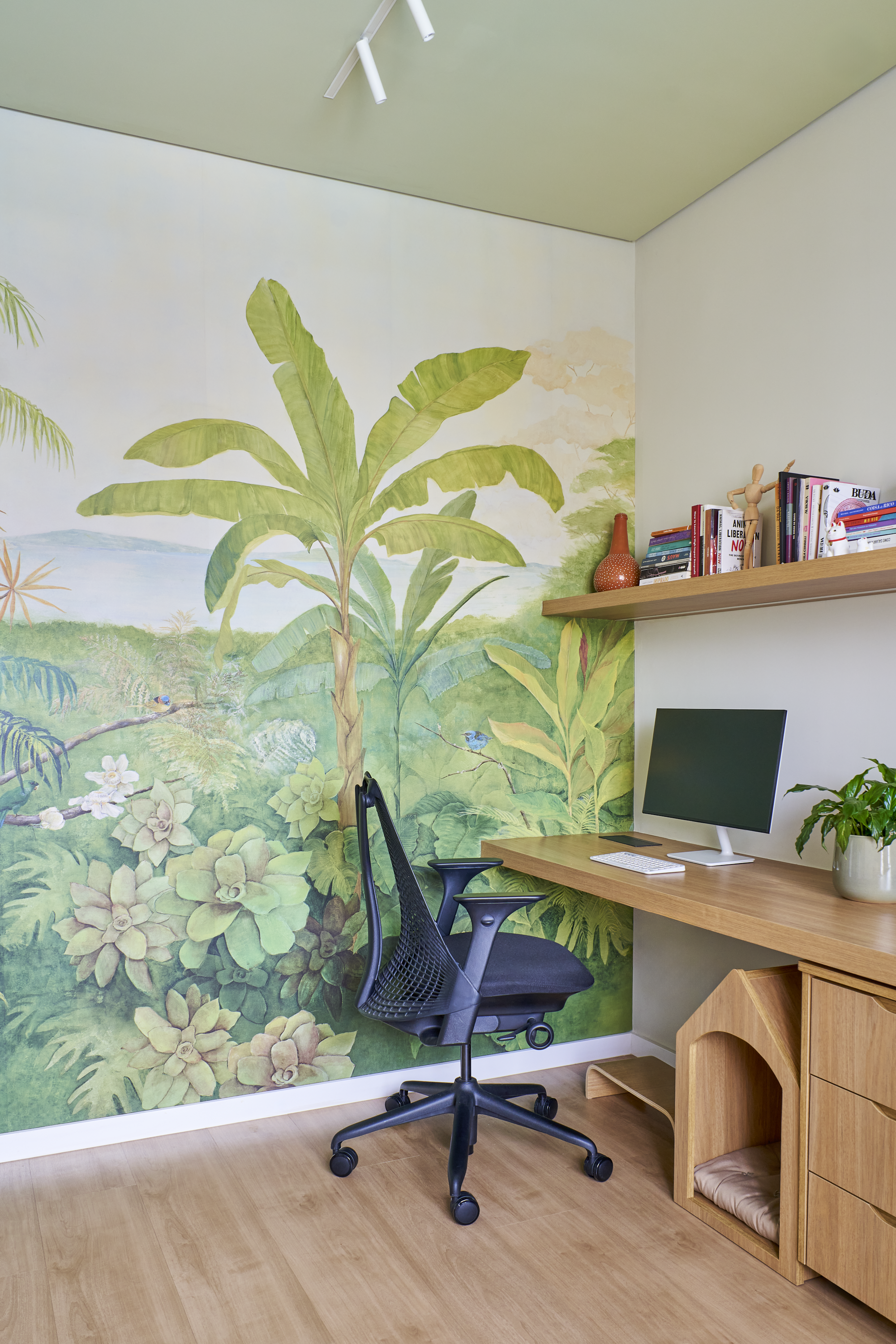 Escritório com parede decorada com mural de vegetação tropical, incluindo árvores e plantas, com uma mesa de escritório, cadeira de escritório, computador, prateleira com livros, vaso de cerâmica, e uma planta em vaso