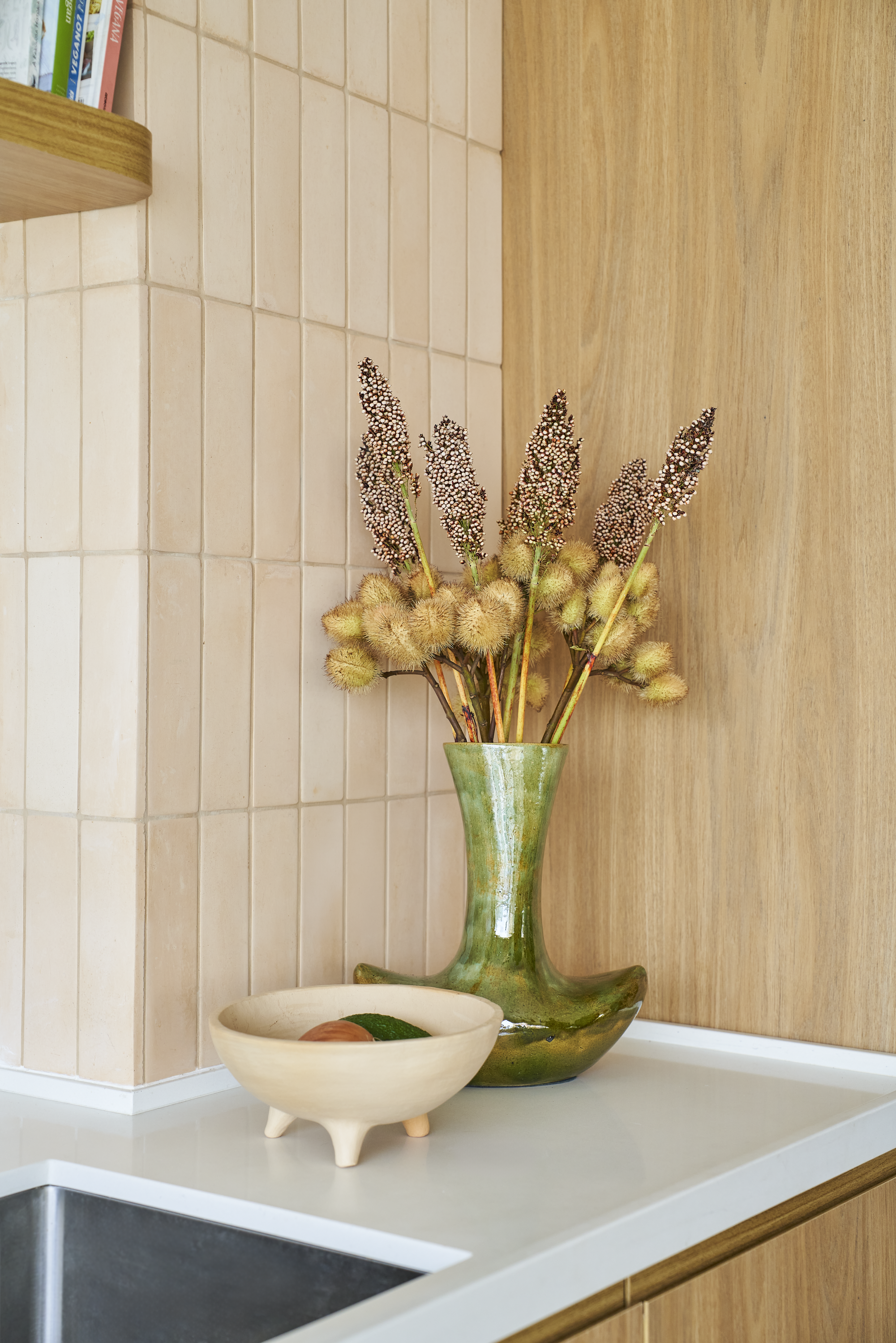 Vaso de cerâmica verde com flores secas, incluindo ramos de flores pequenas rosas e amareladas, sobre bancada de cozinha com tampo branco, ao lado de uma tigela decerâmica branca com três patas, contendo uma fruta verde e uma de cor avermelhada.