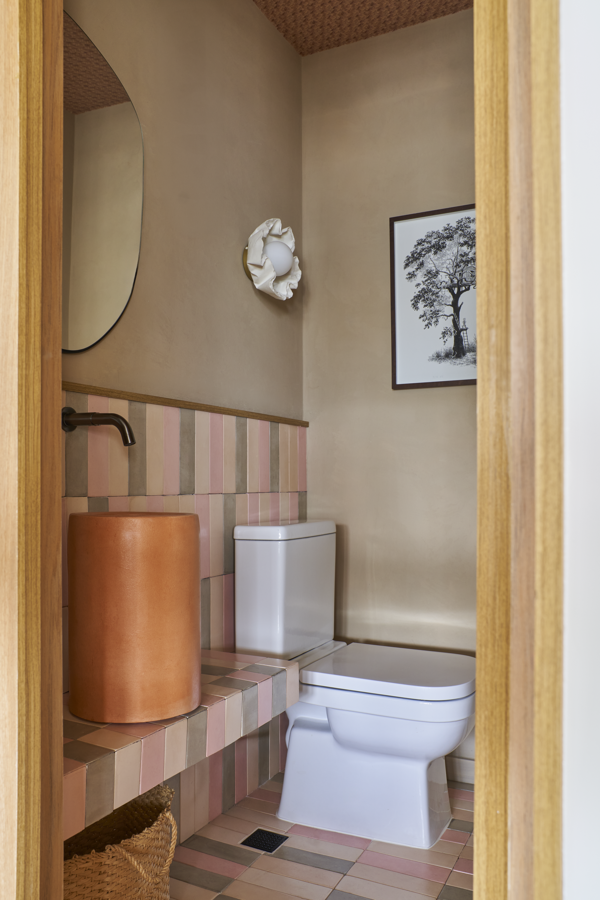 Banheiro com vaso sanitário, espelho no lado esquerdo, parede com azulejos coloridos no meio, parede de tom bege, quadro de árvore na parede direita, luminária decorativa com lâmpada branca e tecido branco no centro