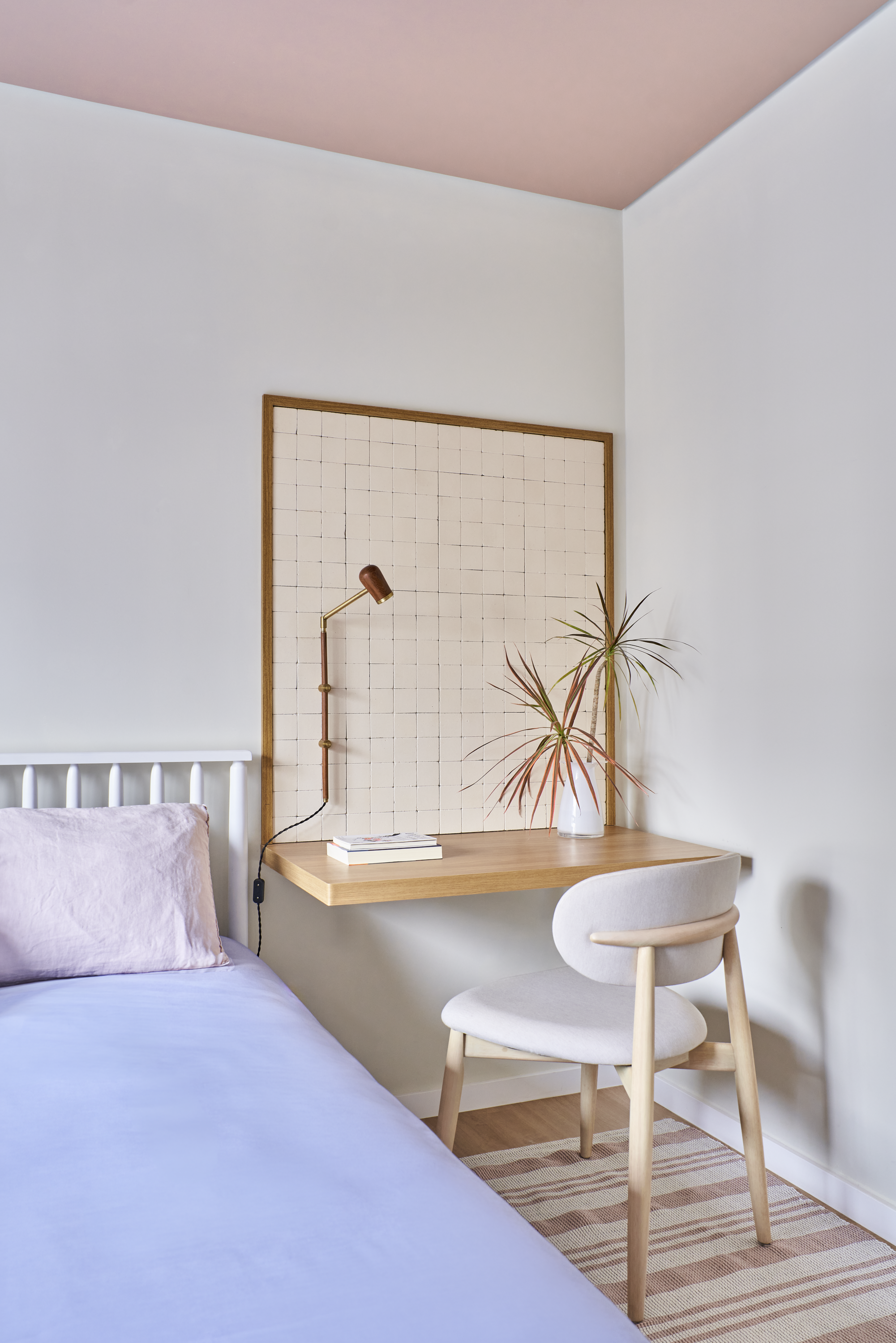 Quarto com cama de lençol azul, travesseiro lilás, mesa de trabalho de madeira com plantas, livros e luminária ajustável, cadeira branca de encosto arredondado, parede de fundo com quadro de malha e parede rosa no teto.