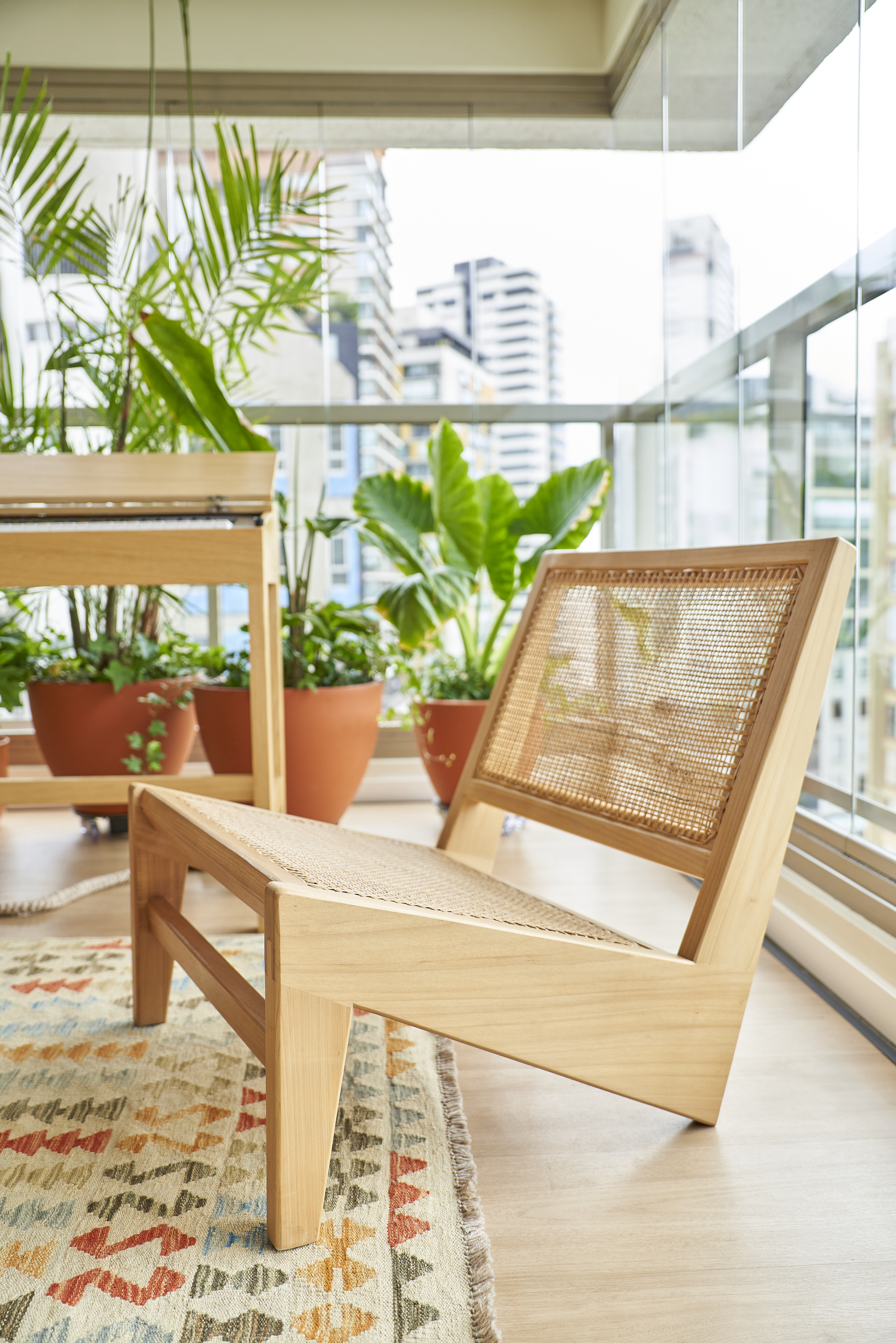 Cadeira de madeira com assento de encosto de cane utilizado em varanda com plantas em vasos de barro e vista de prédios na cidade ao fundo.