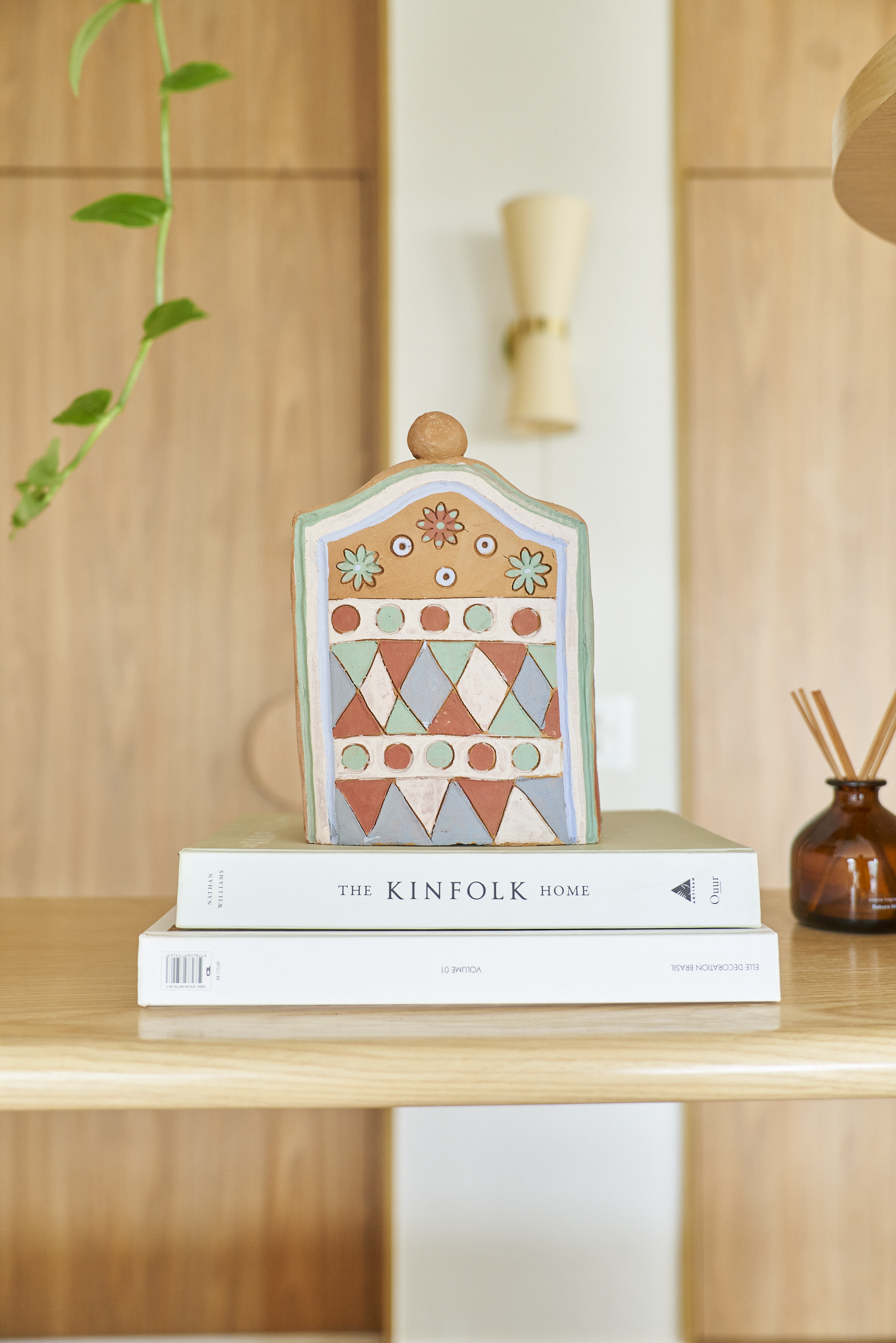 Decoração de interior com uma escultura colorida de redemoinho, apoiada em dois livros, um deles intitulado "The Kinfolk Home", em uma mesa de madeira.
