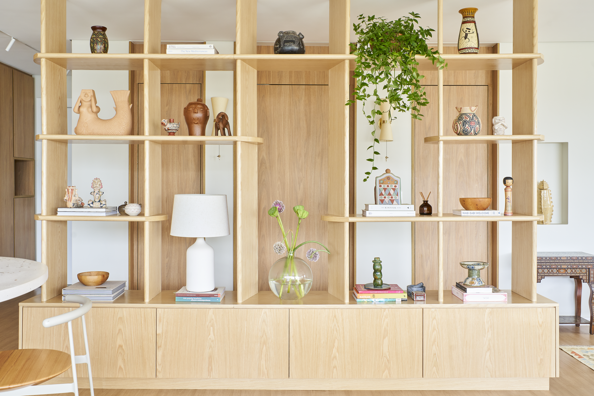 Estante de madeira decorada com vasos, livros, e objetos artesanais, com uma planta pendurada ao lado.