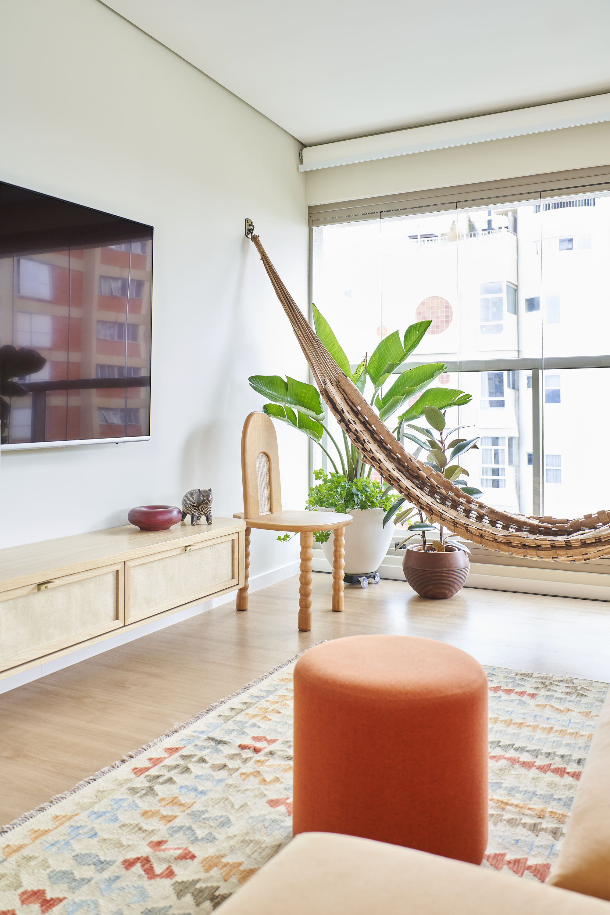 Sala de estar com rede de descanso de palha, plantas grandes na janela, televisão na parede, cadeira de madeira, armário de parede, tapete colorido e um puff laranja.