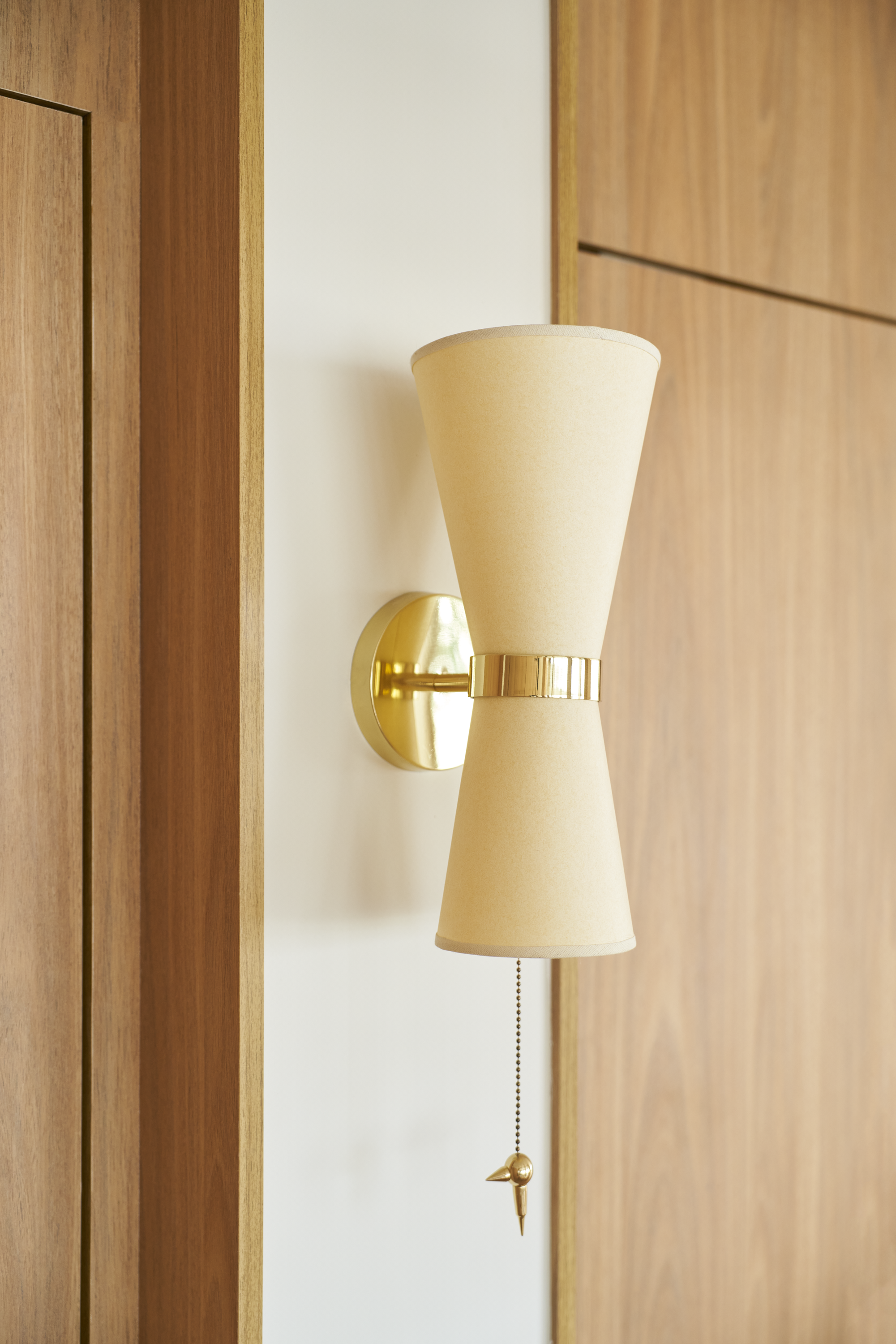Luminária de parede com acabamento dourado e cúpula de tecido bege, presa a uma parede branca com painel de madeira ao fundo.