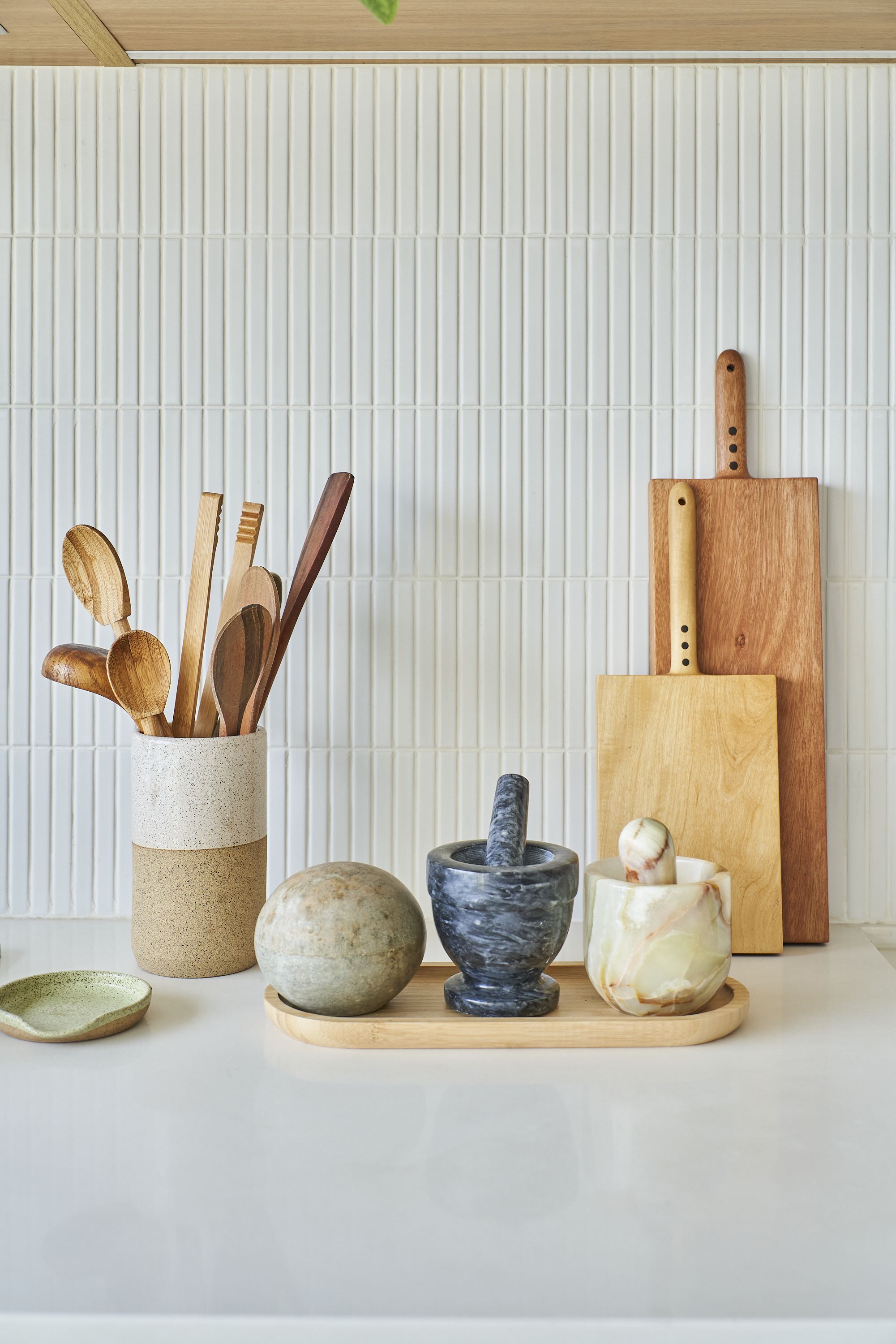 Utensílios de cozinha de madeira, incluindo colheres, tábua de corte e almofariz, dispostos sobre uma bancada de cozinha com parede de azulejos brancos ao fundo.