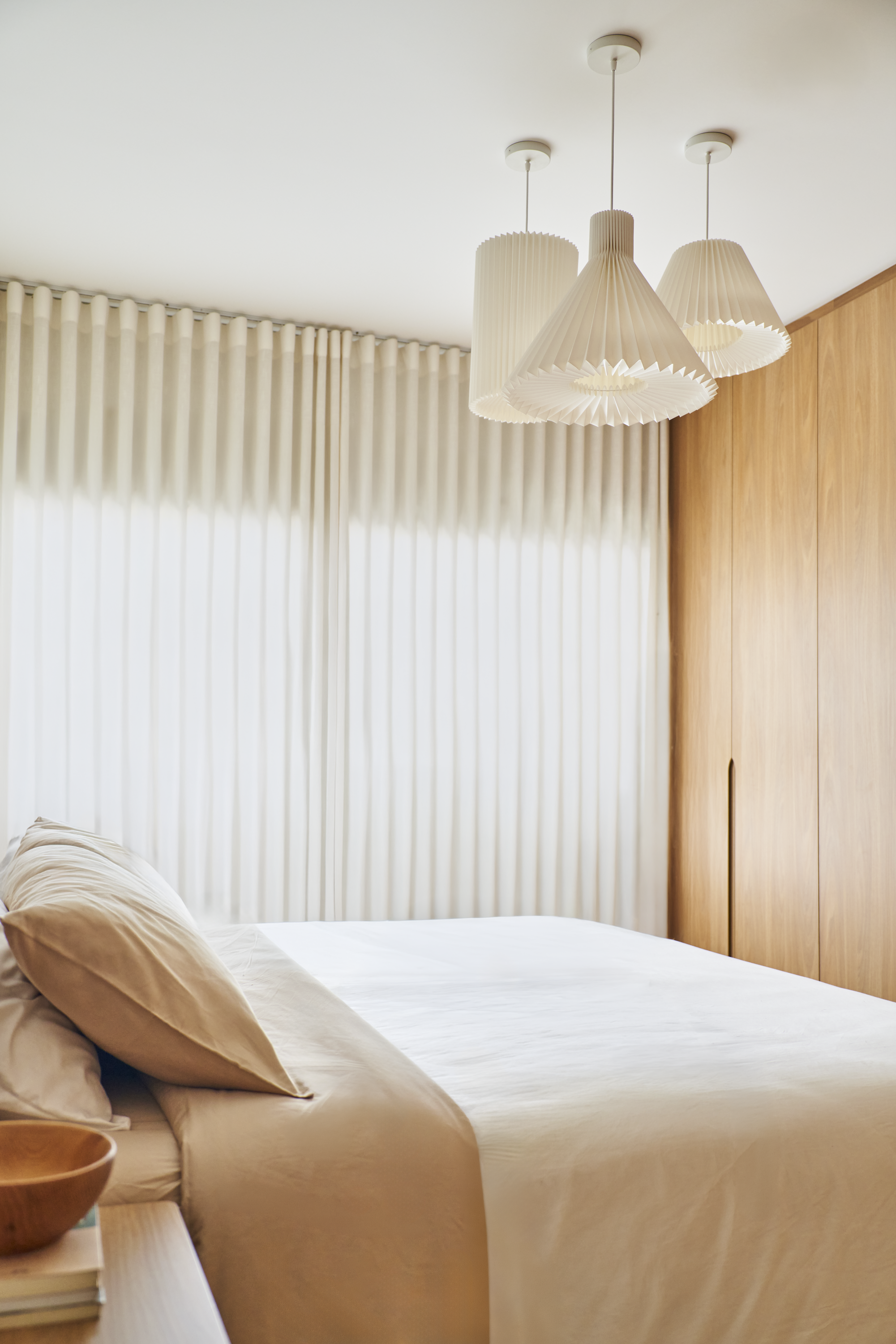 Quarto com paredes de madeira, cortinas brancas, cama com roupas de cama bege, quadros de iluminação pendurados no teto