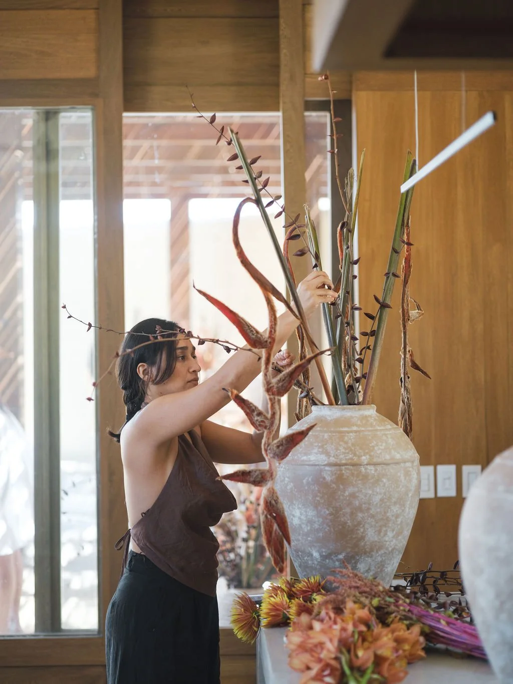 A woman arranging tall dried plants in a large ceramic vase indoors, with a window and wooden interior in the background.