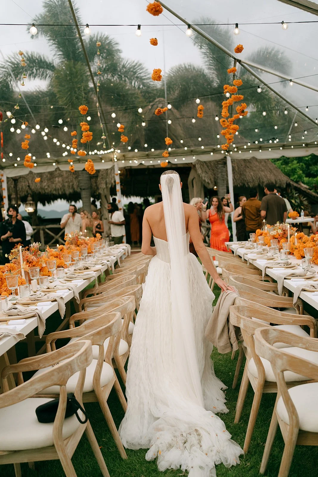 A bride in a white wedding gown with a veil walking through a decorated outdoor reception area. The area has long tables with orange floral centerpieces, surrounded by wooden chairs. String lights and hanging orange flowers decorate the tent ceiling. Guests are mingling in the background.