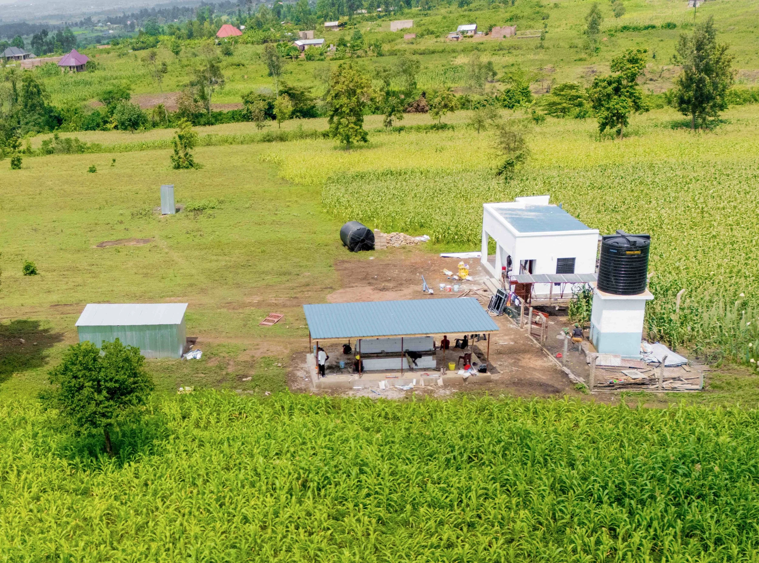 A rural landscape featuring WASH Hub No. 2 with main building, water station, tank, and green fields.