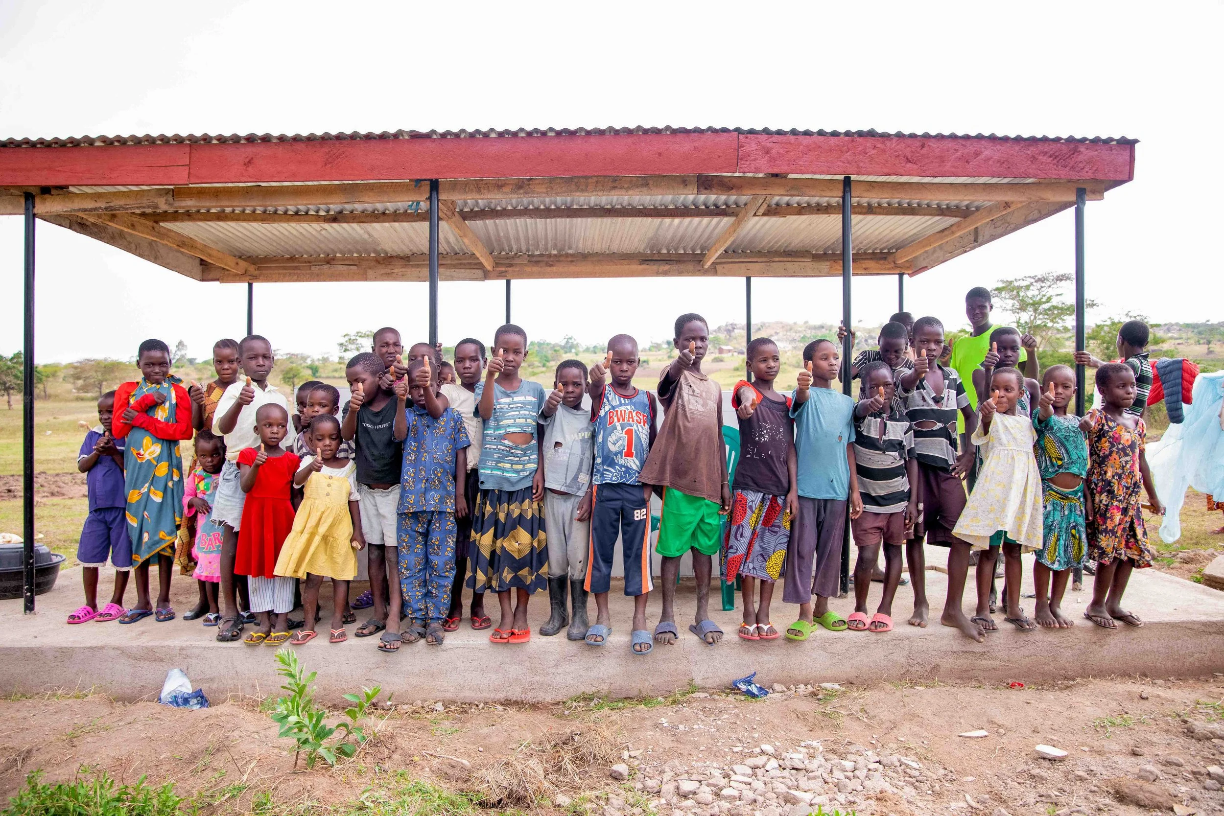 Group of children standing under a shelter, giving thumbs up, in a rural outdoor setting.