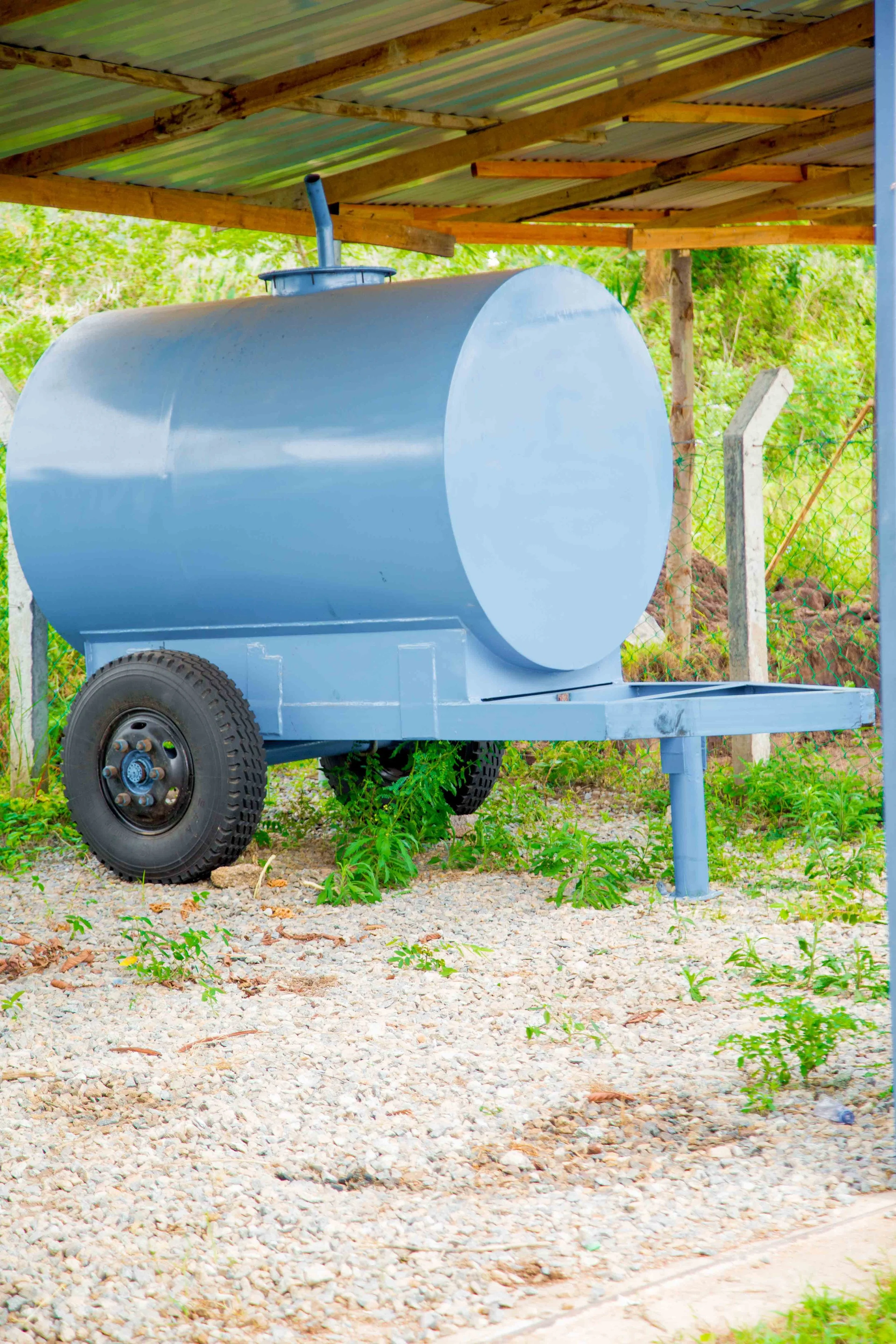 A large, light blue metal water tank on wheels with a pipe on top, situated outdoors under a wooden and metal roof structure, with greenery and a chain-link fence in the background.