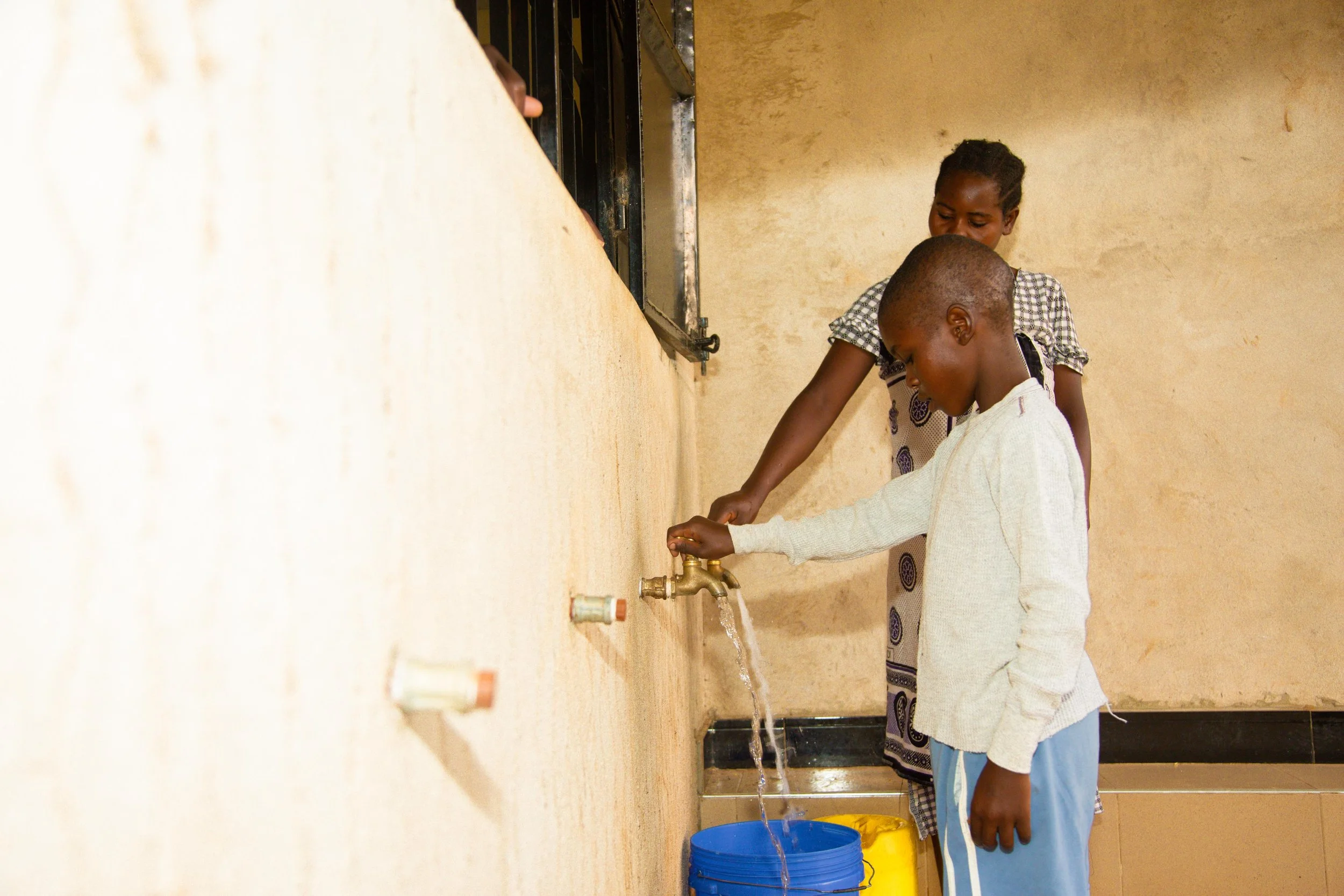 Mother and son standing at a water tap, filling blue and yellow buckets with water.