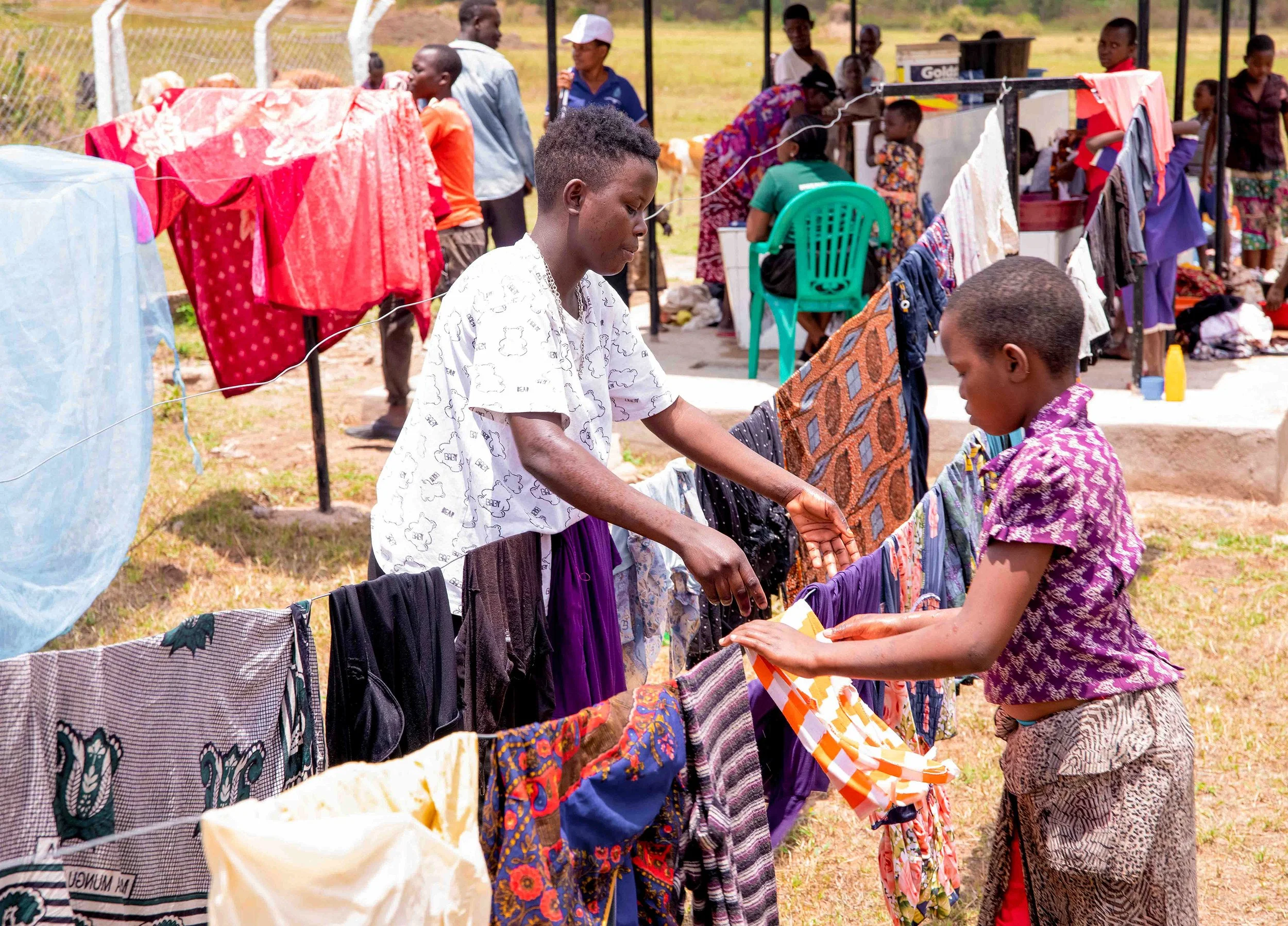 People hanging clothes on a clothesline outside on a sunny day, with several other individuals in the background near a fence and a shaded area.