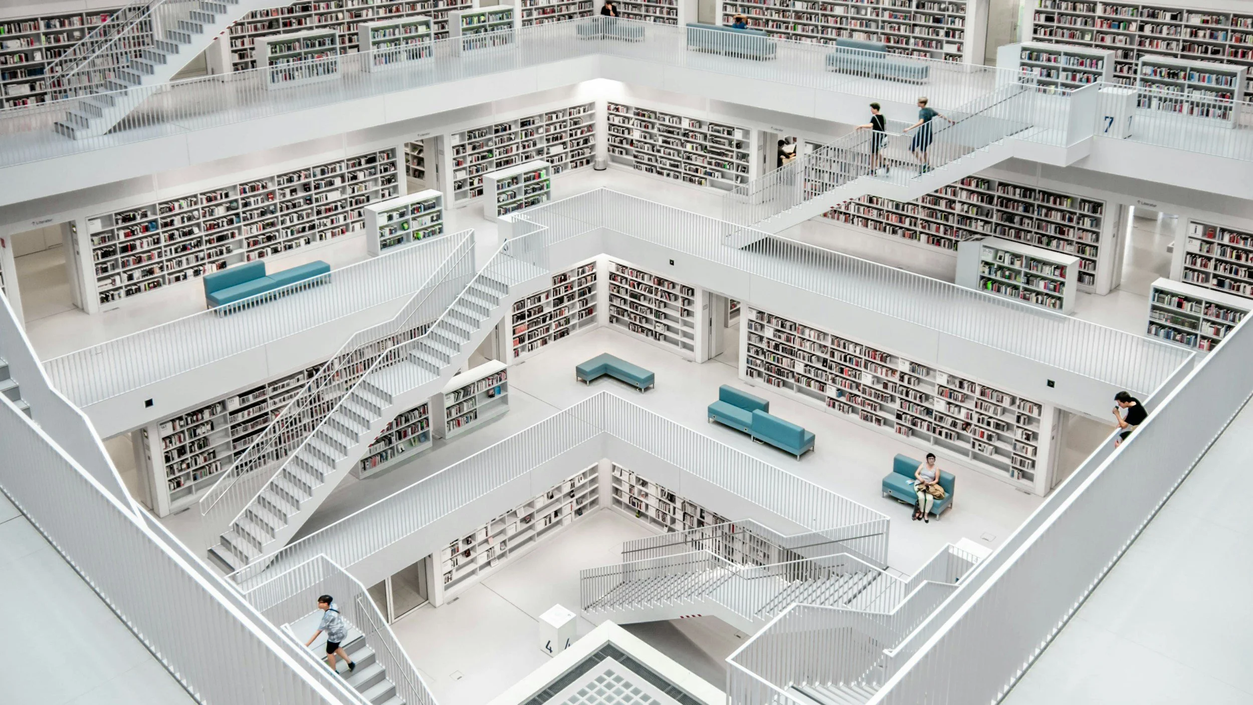 Multi-story modern library with white bookshelves, staircases, and walkways. Few people are browsing or reading books, and there are seating areas with blue benches.