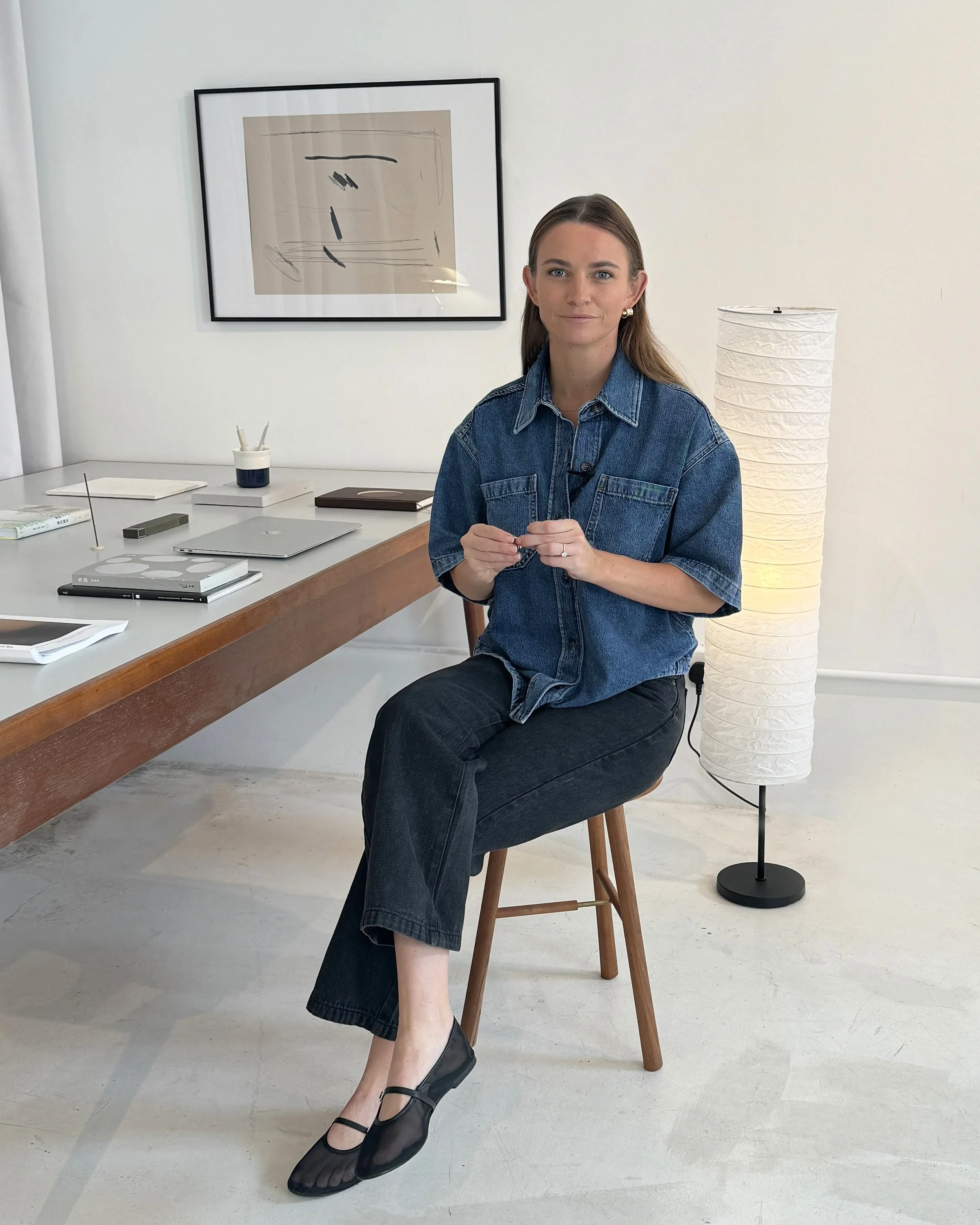 A woman sitting on a wooden chair at a desk in a minimalist office, holding small objects in her hands. She is wearing a denim shirt and black pants, with black shoes. Behind her is a white floor lamp and a framed abstract art piece on the wall.