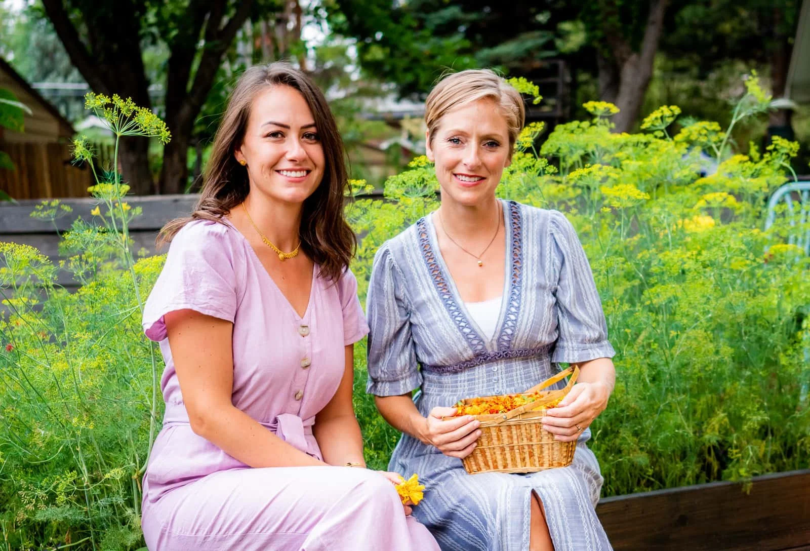 Two women sitting outdoors in a garden, smiling at the camera, surrounded by lush green plants and trees.