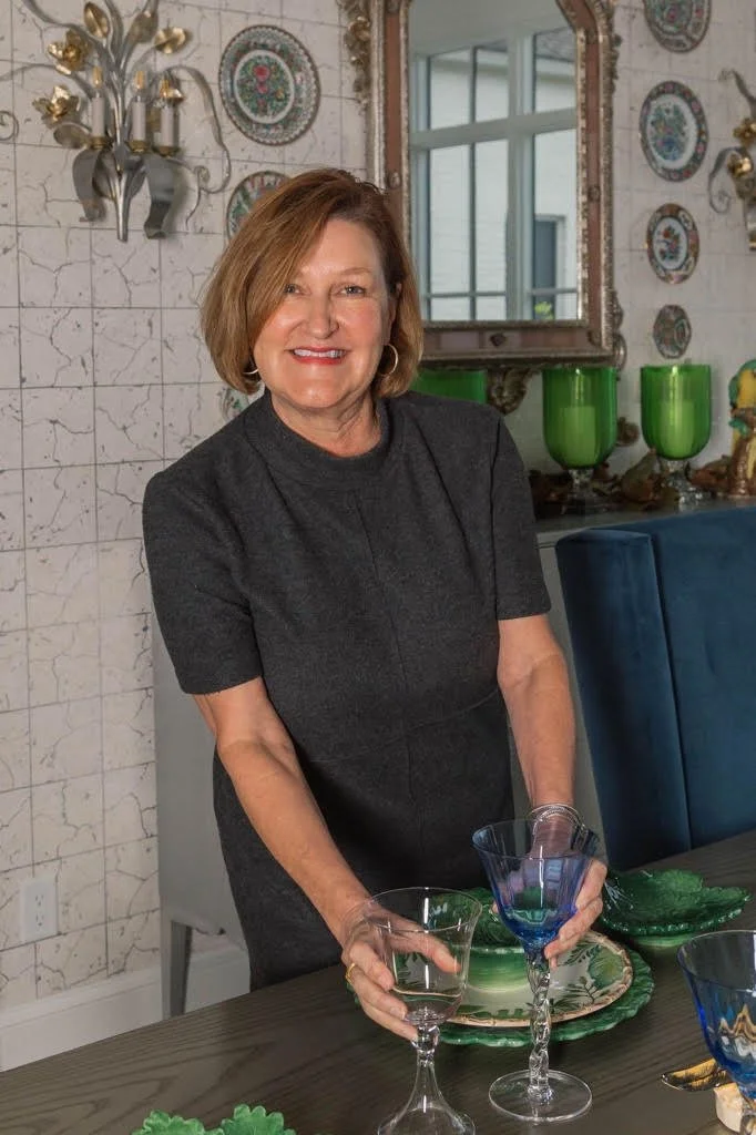 A woman with short brown hair, wearing a dark gray dress, smiling and holding two glasses, standing in a decorated room with a mirror, green vases, and decorative plates on the wall.