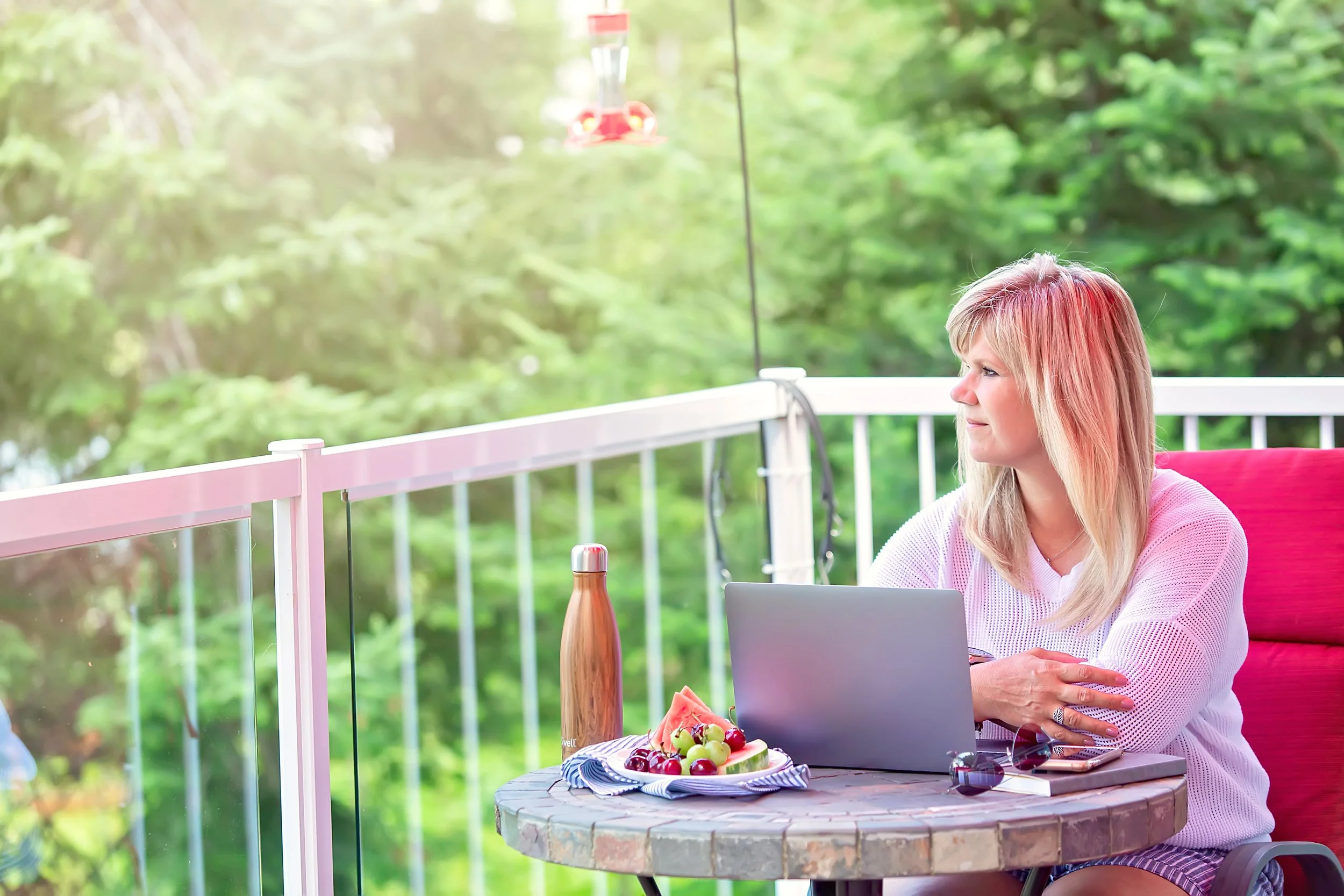 Blonde woman at a table on a deck having breakfast and working on her laptop.