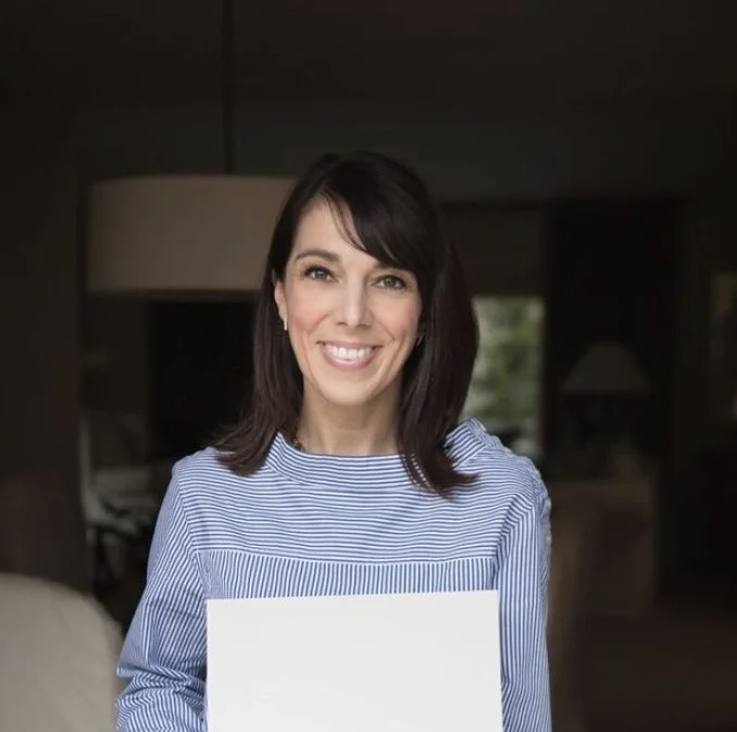 A woman with dark hair and a blue striped shirt smiling and holding a blank white board in a cozy living room.