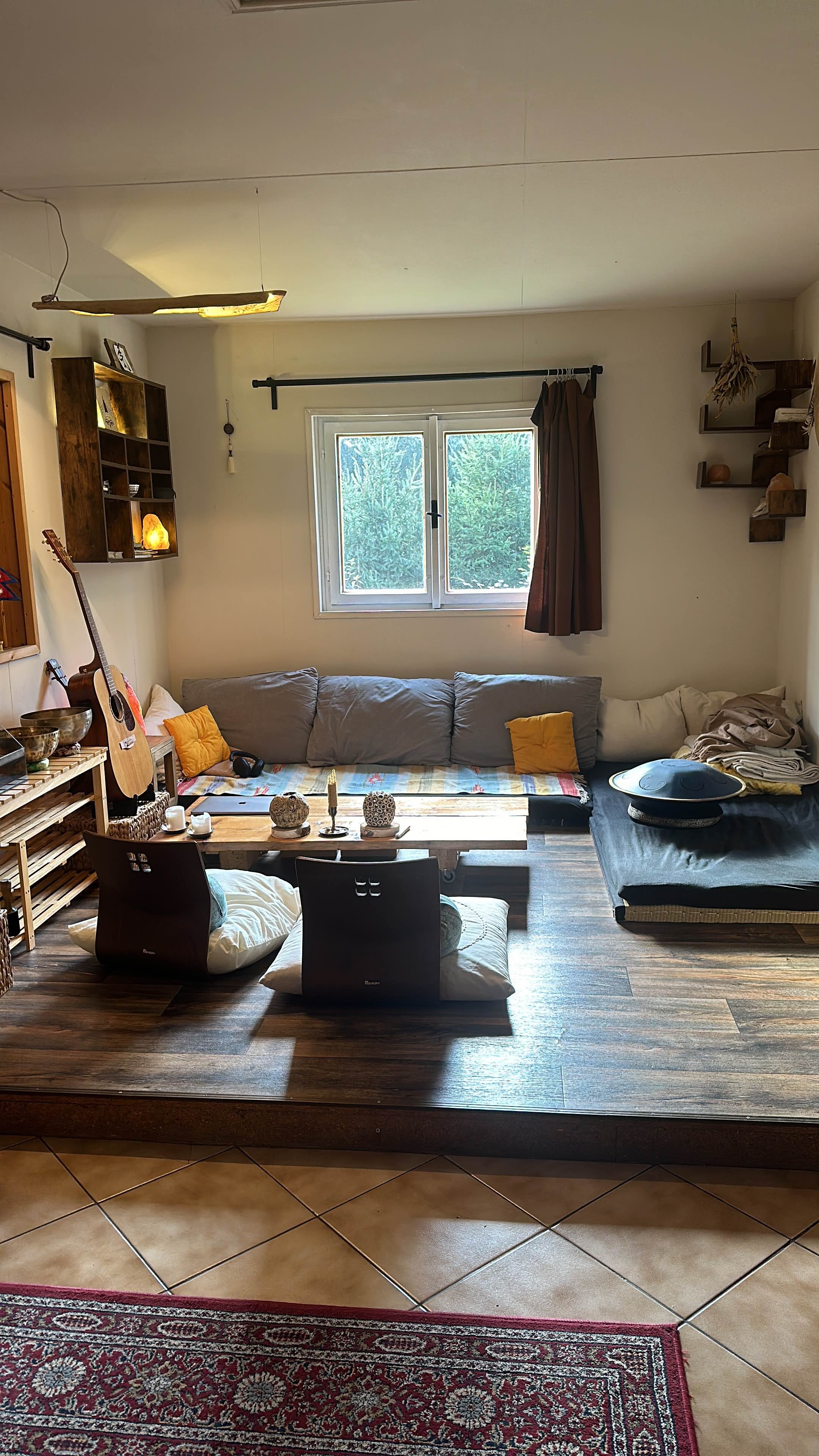 Cozy living room with a low wooden table, cushions, gray sofa with yellow pillows, acoustic guitar, windows, and wall shelves. A handpan and decorative items are on the table.